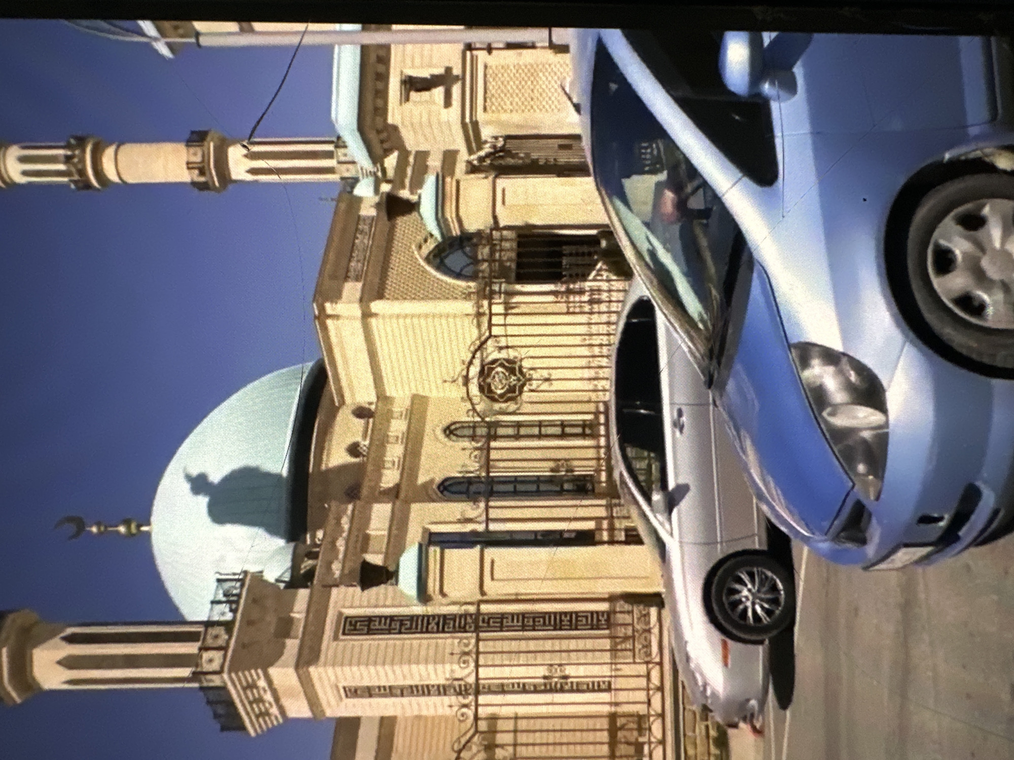 The image captures a street scene in Кызыл-Булак, Kyrgyzstan, featuring a mosque with a large, light blue dome and minarets, constructed from light-colored brick. A crescent moon symbol is visible atop one of the minarets. In the foreground, two cars are parked on a paved surface. The car on the left is silver, while the car on the right is light blue. A decorative metal fence with an intricate design separates the mosque from the street. The sky is clear and blue.