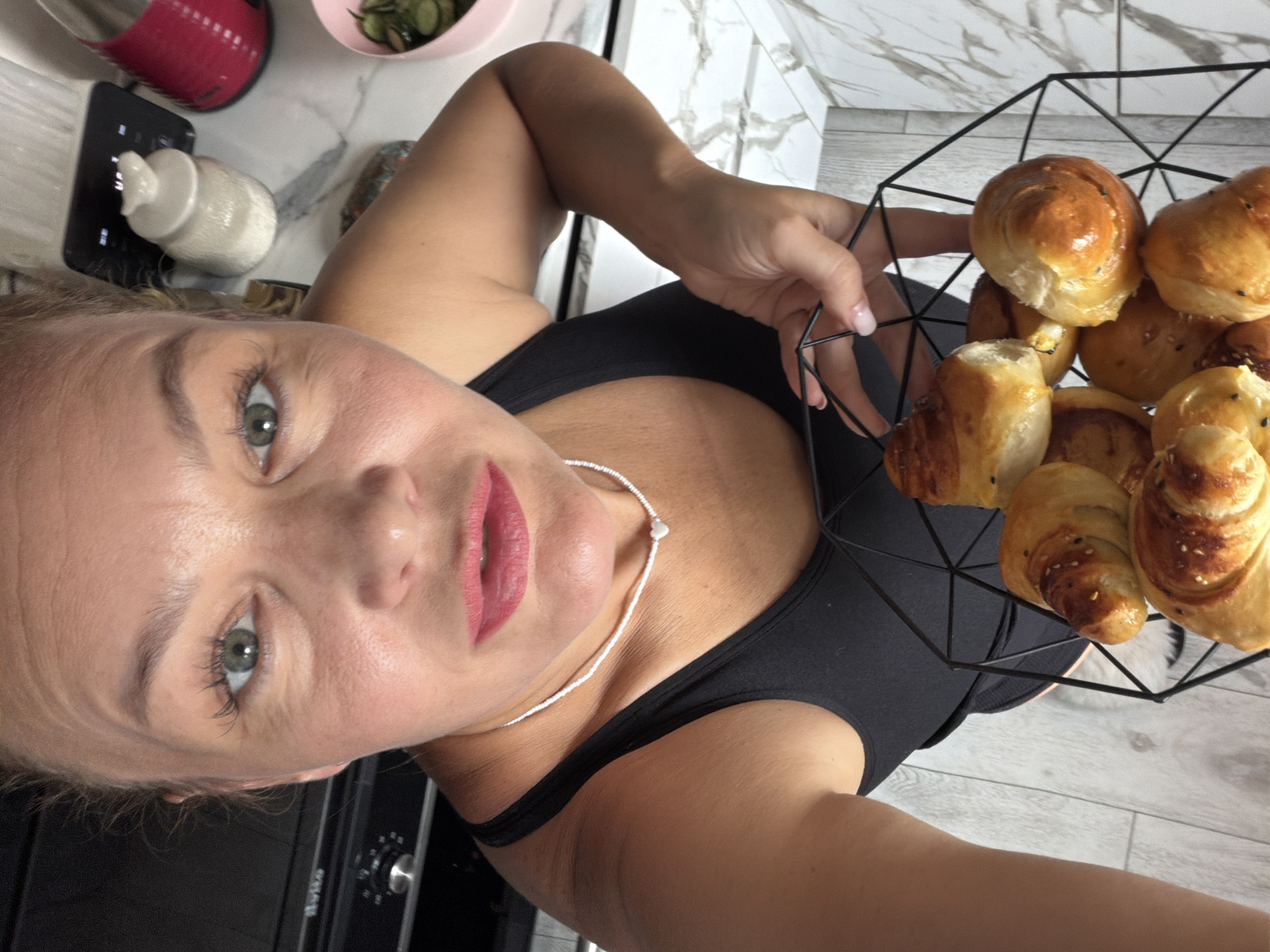 A woman with blonde hair pulled back, wearing a black tank top and a thin white necklace with a heart pendant, holds a black wire basket filled with golden-brown baked goods, possibly croissants or rolls, topped with sesame seeds. She is in a kitchen with light-colored countertops and light-colored wood-patterned flooring. In the background, on the counter, is a black kitchen appliance with a dial, a white ceramic jar with a bird-shaped lid, and a pink electric kettle. A light pink bowl containing sliced green vegetables is also visible. The woman is looking directly at the camera, her expression neutral. The scene is indoors, appearing to be a domestic kitchen environment, not specifically identifiable as being in the city of Chaikovsky, Russia.