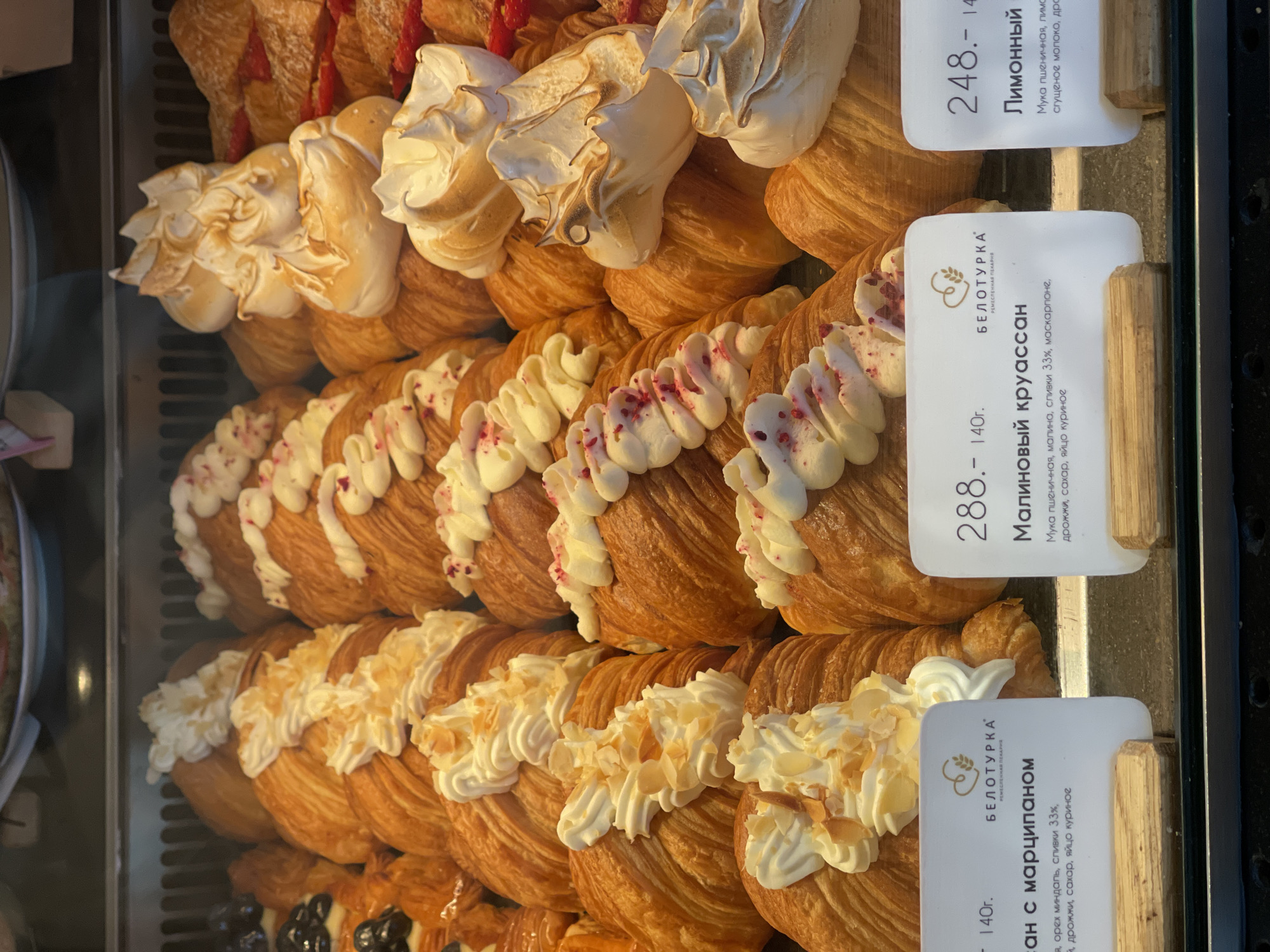 The image shows a display case of pastries in Izhevsk, Russia. Croissants are the main subject, topped with various creams and toppings: a row of toasted meringue, a row with white cream and red sprinkles, and a row with white cream and sliced almonds. Price tags provide information in Russian. One tag reads "Малиновый круассан" (raspberry croissant), listing ingredients like wheat flour, raspberries, 33% cream, and mascarpone. Another reads "ан с марципаном" (with marzipan), listing ingredients like almonds, 33% cream, sugar, and eggs. A third tag reads "Лимонный" (Lemon), listing ingredients like wheat flour, lemon, and condensed milk. The word "БЕЛОТУРКА" (Beloturka) is also prominently featured on the price tags, likely the name of the bakery. Prices are listed in rubles, and the weight of each pastry is listed as 140 grams. The lighting is bright, highlighting the pastries' golden-brown crusts and the creamy toppings.