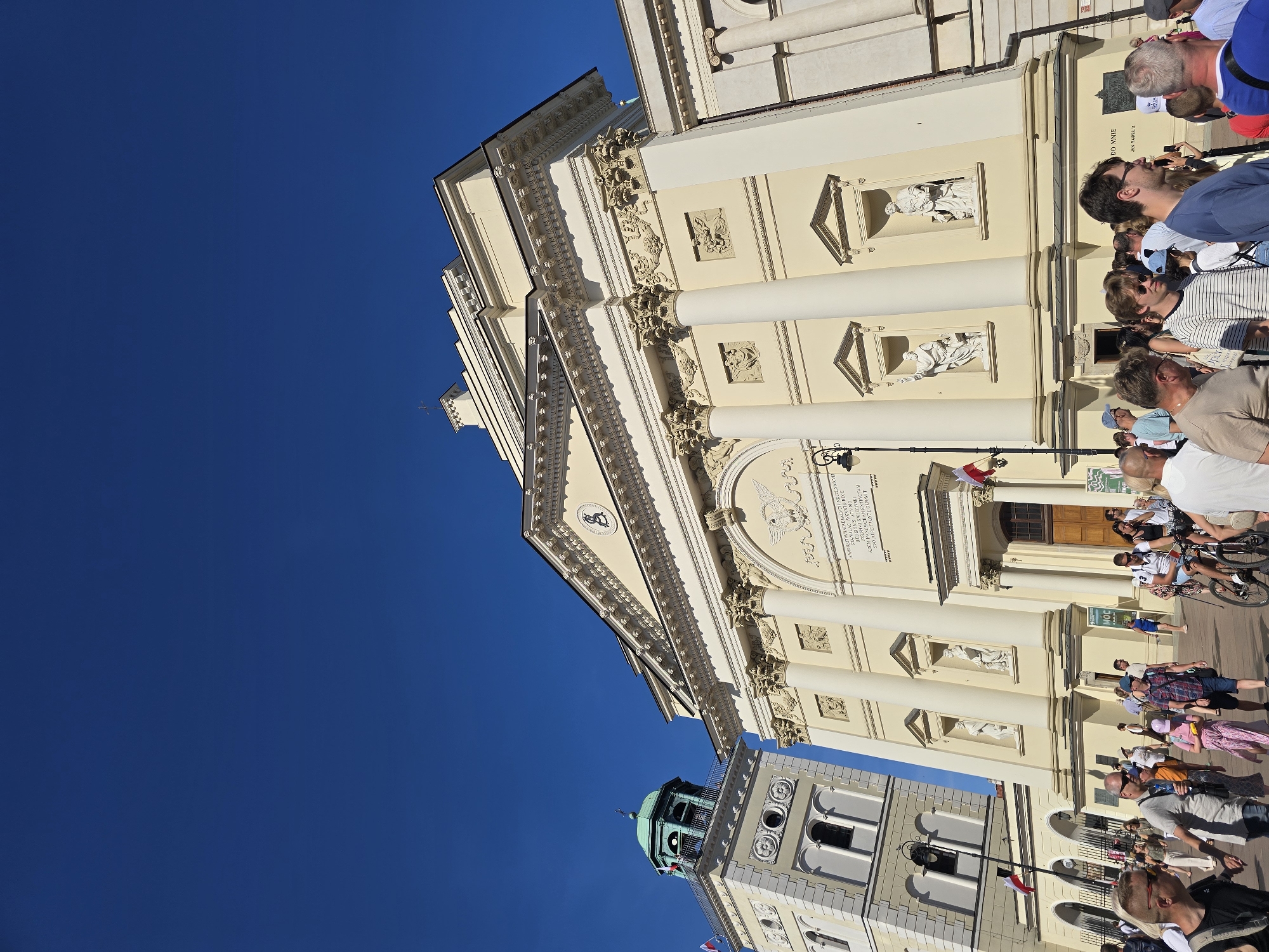 In this medium-shot photograph taken on a bright, clear day in Warsaw, Poland, a crowd of people is gathered in front of a grand, pale yellow neoclassical building. The building features large white columns, ornate architectural details, and a prominent pediment with a coat of arms. To the left of the main building is a taller, lighter-colored tower with a green dome, also featuring architectural ornamentation and a flagpole with a red and white flag. The sky above is a deep, cloudless blue.
The foreground is populated by a diverse group of individuals, varying in age and attire. Many are walking, while some are stationary, observing the scene or engaging in conversation. A man on a bicycle is visible on the right side of the frame, suggesting a lively public space. The people are dressed in casual, warm-weather clothing. The overall atmosphere appears to be one of tourism or a public gathering. The architecture strongly suggests the building is a church or significant public edifice. The presence of Polish flags hints at the location within Warsaw. Based on the architectural style and the likely presence of St. John's Archcathedral and its bell tower in Warsaw's Old Town, this scene is likely depicts Castle Square (Plac Zamkowy).
