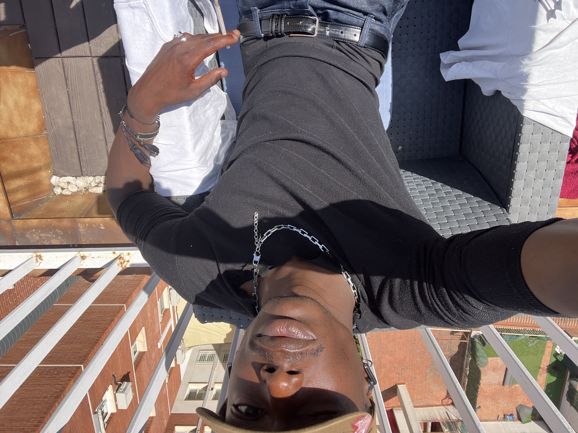 An upside-down, eye-level shot captures a person of color taking a selfie on a sunny balcony or terrace in Barcelona, Spain. The individual is positioned horizontally across the frame, facing upwards, with their head at the bottom and feet at the top of the image.
The subject is wearing a black long-sleeved top and dark blue jeans with a black leather belt. A silver-colored chain necklace with large links hangs around their neck. On their left wrist, multiple bracelets are visible, including braided dark ones and one with tassels. A silver ring adorns the ring finger of their left hand, which is resting near their torso. A light-colored (possibly white) t-shirt is draped over the chair next to them. A khaki or brown cap is worn backwards, partially covering the top of their head. The person has dark skin, full lips, and is looking directly towards the camera with a neutral expression.
They are reclining on a dark grey, woven outdoor chair. In the background, beyond a white metal railing, is a cityscape under bright sunlight. Buildings with red brick facades and some lighter-colored structures are visible, featuring windows and outdoor air conditioning units. Terracotta roof tiles are also seen on some buildings, and a patch of artificial green grass is visible on a rooftop. Strong shadows indicate a bright, clear day, likely late morning or early afternoon. The overall impression is of a relaxed moment outdoors in a vibrant urban environment. There is no visible text.
