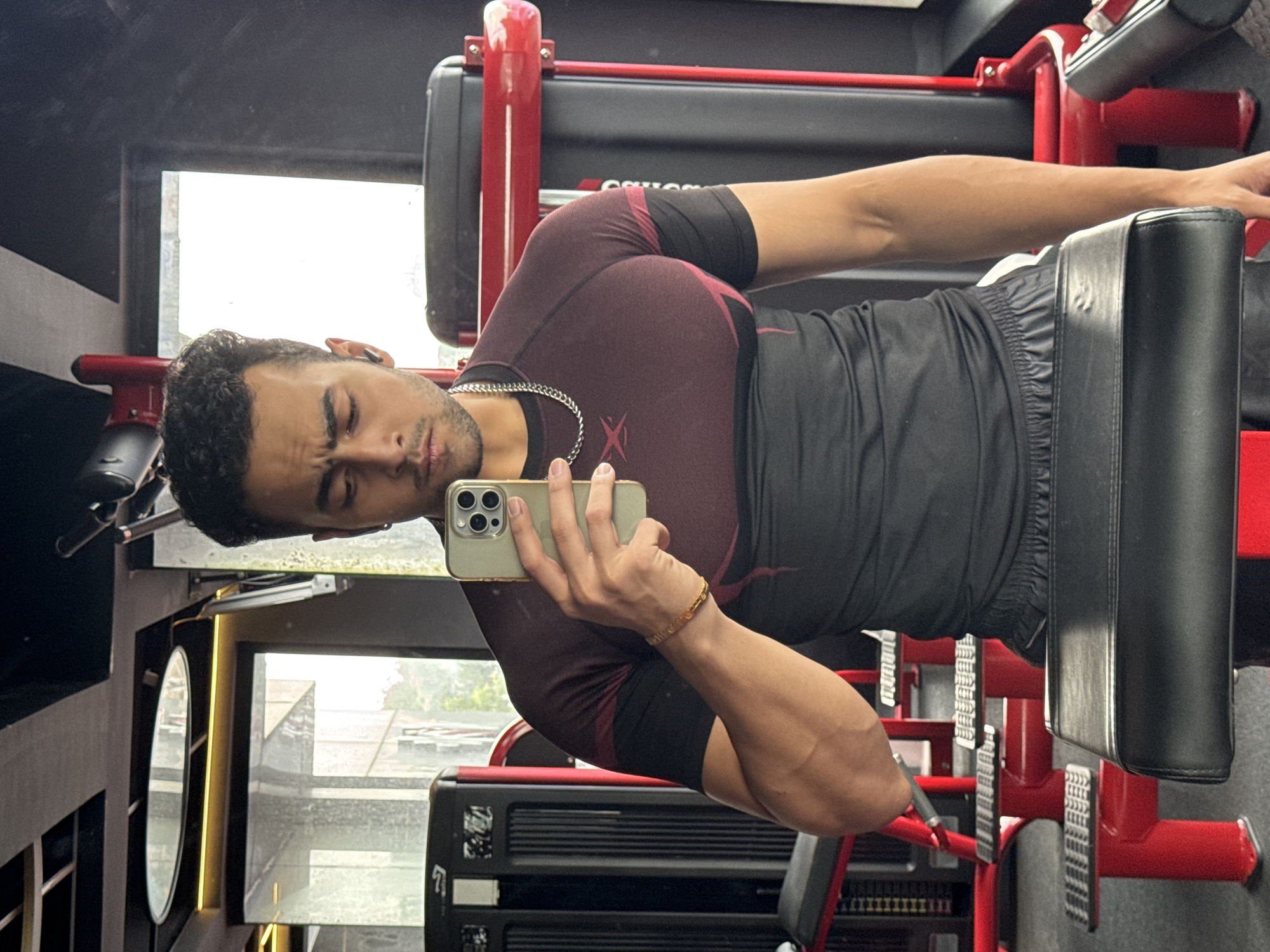A young South Asian man with curly dark hair and a beard takes a mirror selfie in a gym. He is wearing a maroon and black t-shirt and black shorts. He is holding a gold-colored smartphone in his right hand and has a silver chain around his neck and a gold bracelet on his left wrist. The gym is equipped with red and black weight machines and has large windows with a view of buildings. The environment suggests the photo was taken during daylight hours. The details provided in the image do not offer specific information about the location being Kolkata, India.