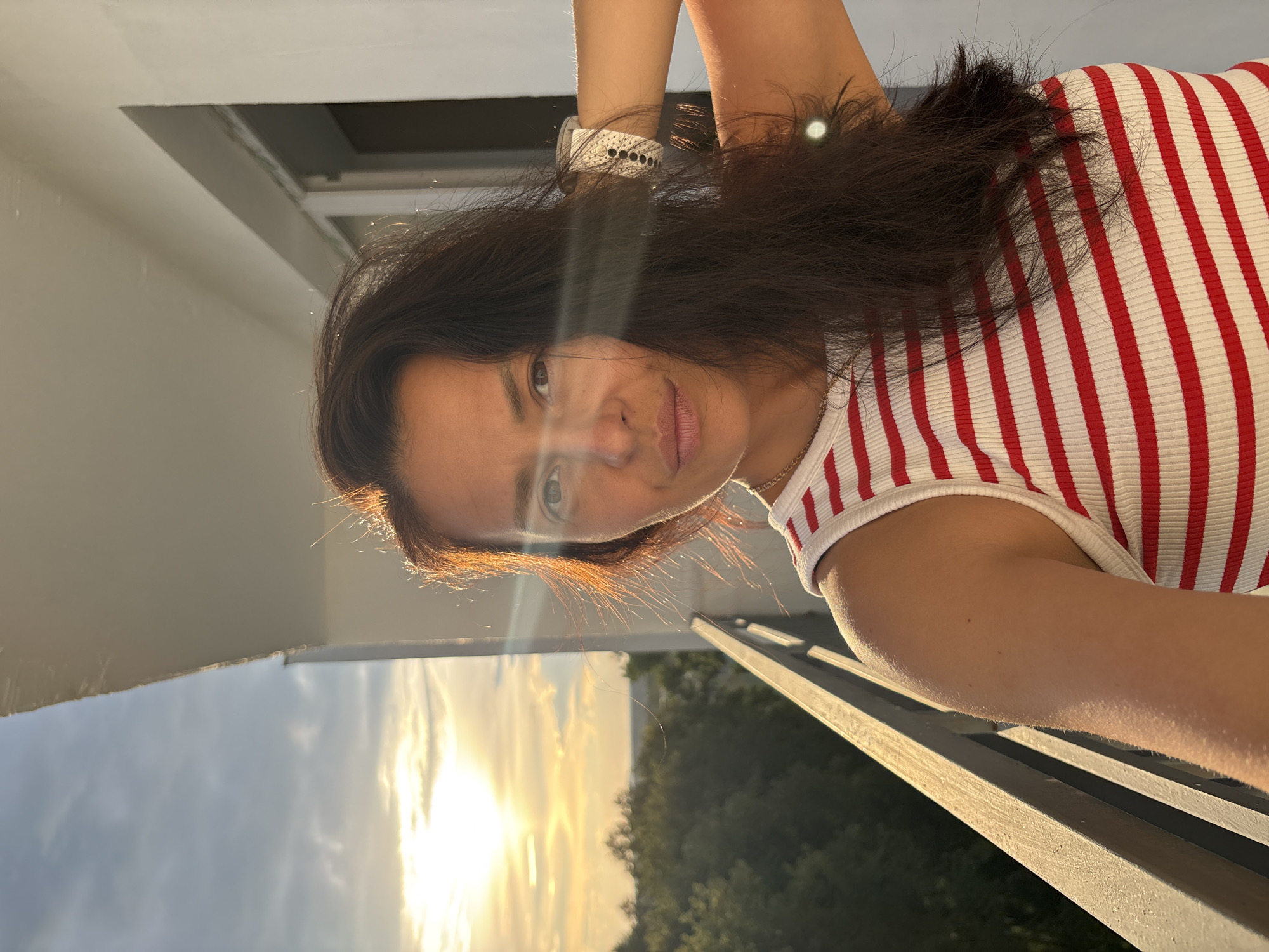 The image shows a woman taking a selfie on a balcony with a sunset in the background. She has long, dark hair and is wearing a red and white striped shirt. A white smartwatch is visible on her left wrist. The sun is setting, casting a warm golden light on the scene. The sky is filled with clouds, and the light suggests it is either sunrise or sunset. The setting appears to be a residential building, possibly an apartment complex, given the balcony railing and the view of trees and sky. The woman's expression is neutral. There is no visible text in the image.