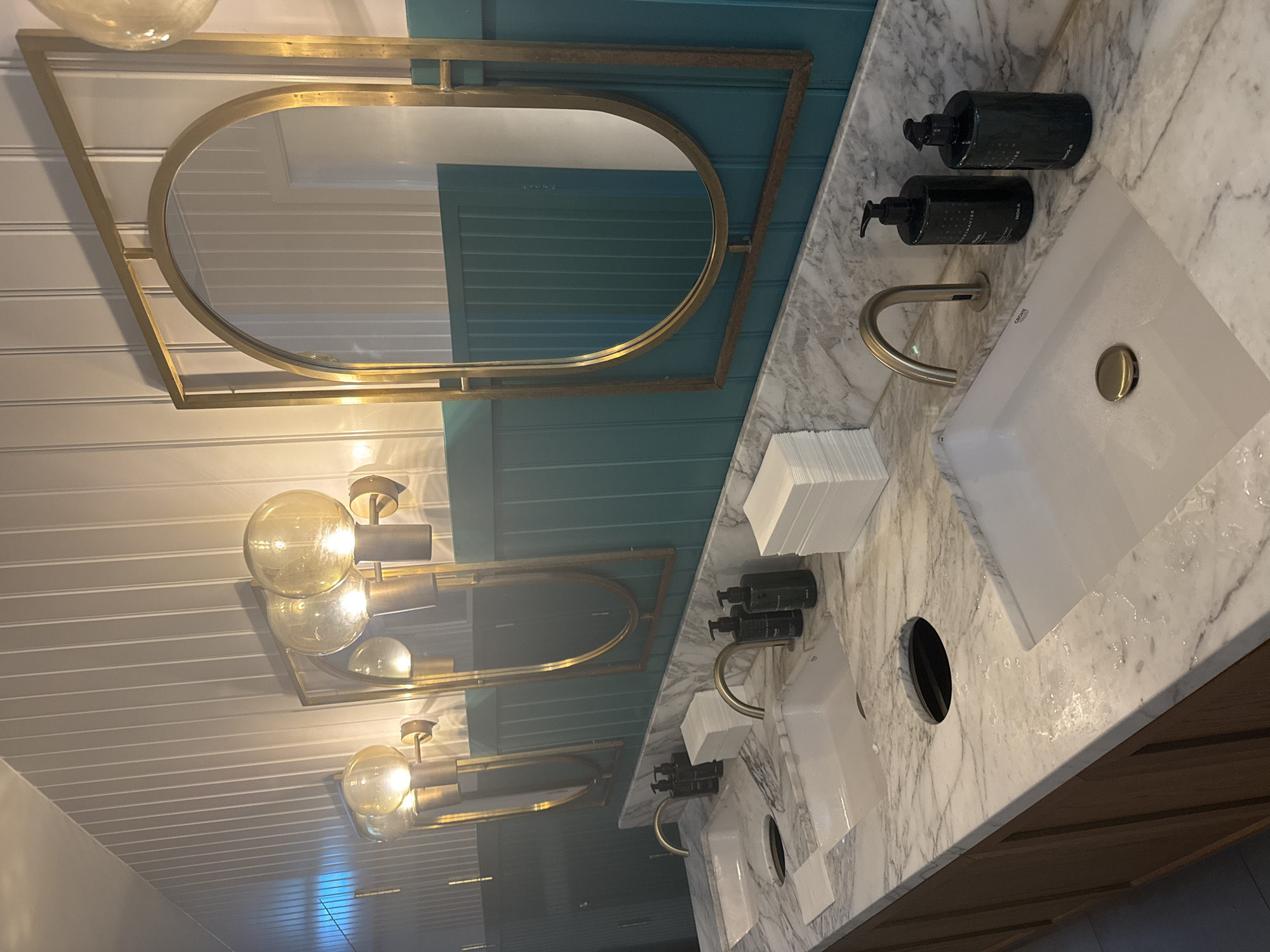 This is a photograph of a modern public restroom with a row of sinks. The countertops are made of white marble with gray veining. Each sink is a white ceramic basin with a brushed nickel faucet. To the right of each faucet are black pump bottles of hand soap. To the right of the soap bottles are stacks of white paper towels. Above the sinks are large, gold-framed mirrors with an arched top. Wall-mounted light fixtures with large, globe-shaped bulbs are positioned above and between the mirrors. The walls behind the mirrors are paneled in a teal green color, and the upper walls are paneled in white. The image appears to be taken in a well-lit, clean restroom. It is not possible to determine the specific location in Copenhagen, Denmark from the image alone.