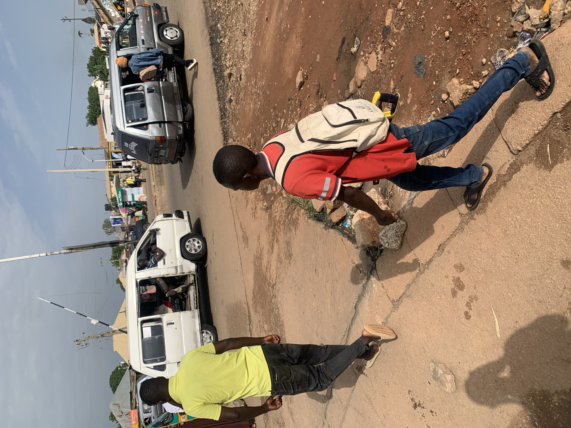 The image depicts a street scene in Gura Top, Nigeria, with two young men walking on a paved sidewalk. A white minivan, likely a public transport vehicle, is stopped on the road to the left, with its side door open and passengers visible inside. To the right of the white minivan, a silver minivan is also on the road, with its rear door open and a young woman in a denim jacket and orange beanie exiting or entering, carrying a bag. The ground is a mix of cracked concrete sidewalk and reddish-brown dirt road, with scattered debris including rocks and plastic. In the background, there are buildings, utility poles, and other vehicles, suggesting a bustling urban environment. The lighting indicates it is daytime, likely late afternoon, with some shadows cast on the ground. The overall scene conveys a sense of everyday activity in a Nigerian city.