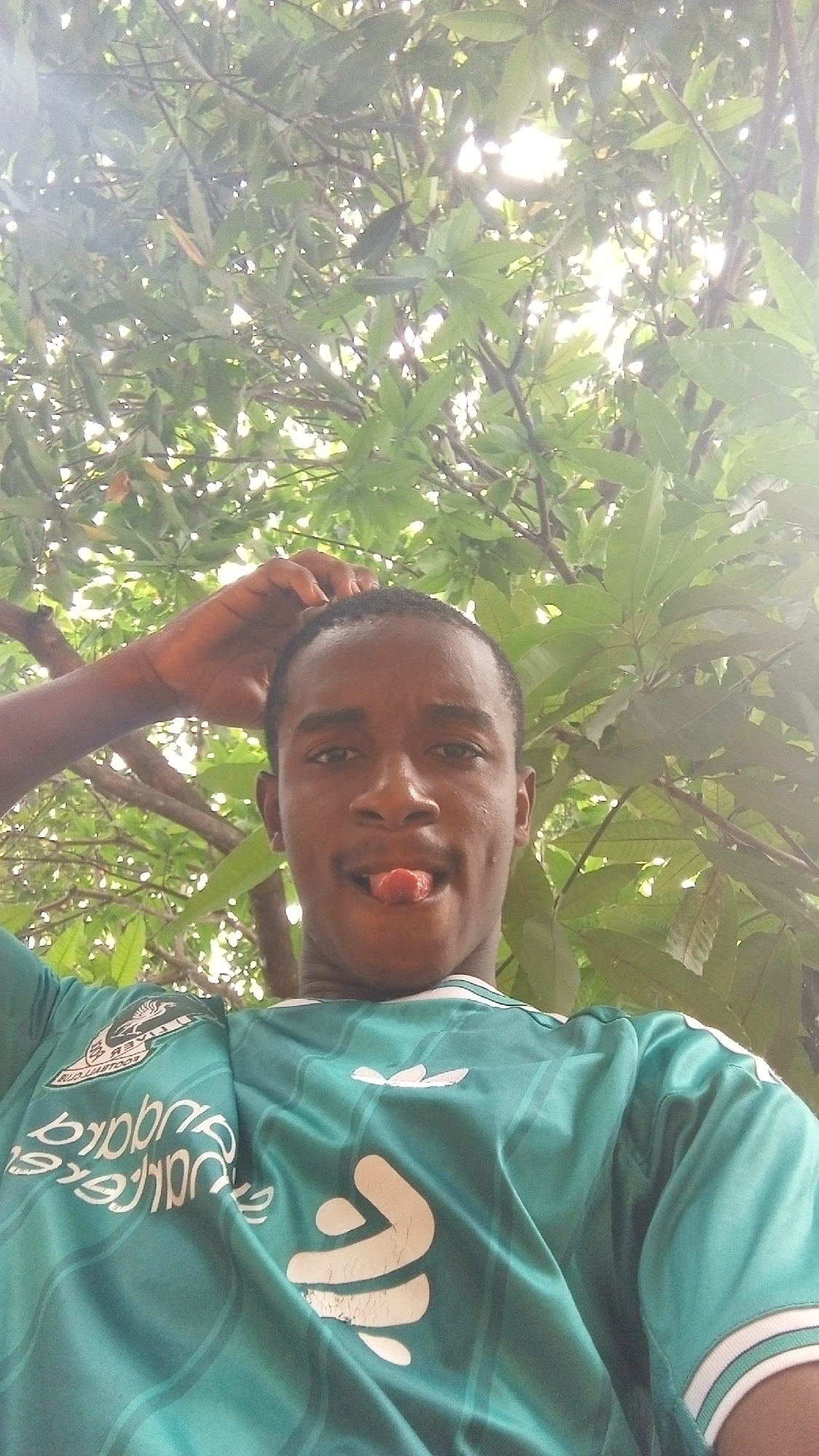 A medium shot taken from a low angle shows a young Black man with short, dark hair and brown eyes. He is sticking his tongue out and has his right hand on his head. He is wearing a teal, white, and green soccer jersey with an Adidas logo on the right shoulder. Behind him is dense green foliage and tree branches obscuring the sky. The image was apparently taken outdoors. The city is not discernible in the image.
