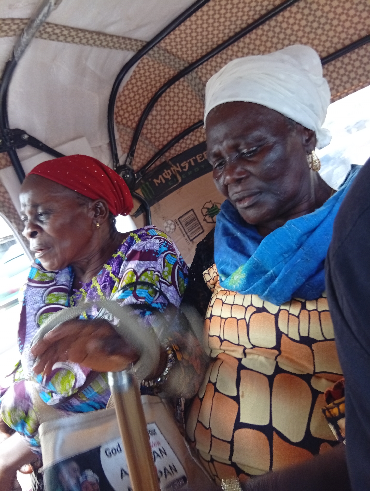 The image shows two elderly women sitting in the back of a motorized tricycle, commonly known as an "okada," in the city of Bukuru, Nigeria. The woman on the left is wearing a red headscarf and a colorful patterned dress, while the woman on the right is wearing a white headscarf, a blue scarf, and a yellow and black patterned dress. Both women appear to be looking downwards, and their expressions are neutral. The okada has a canopy overhead with a brown patterned fabric, and the background is blurred, suggesting movement. A cardboard box with the "MONSTER ENERGY" logo is visible behind the women. The overall scene depicts everyday transportation in an urban Nigerian setting.