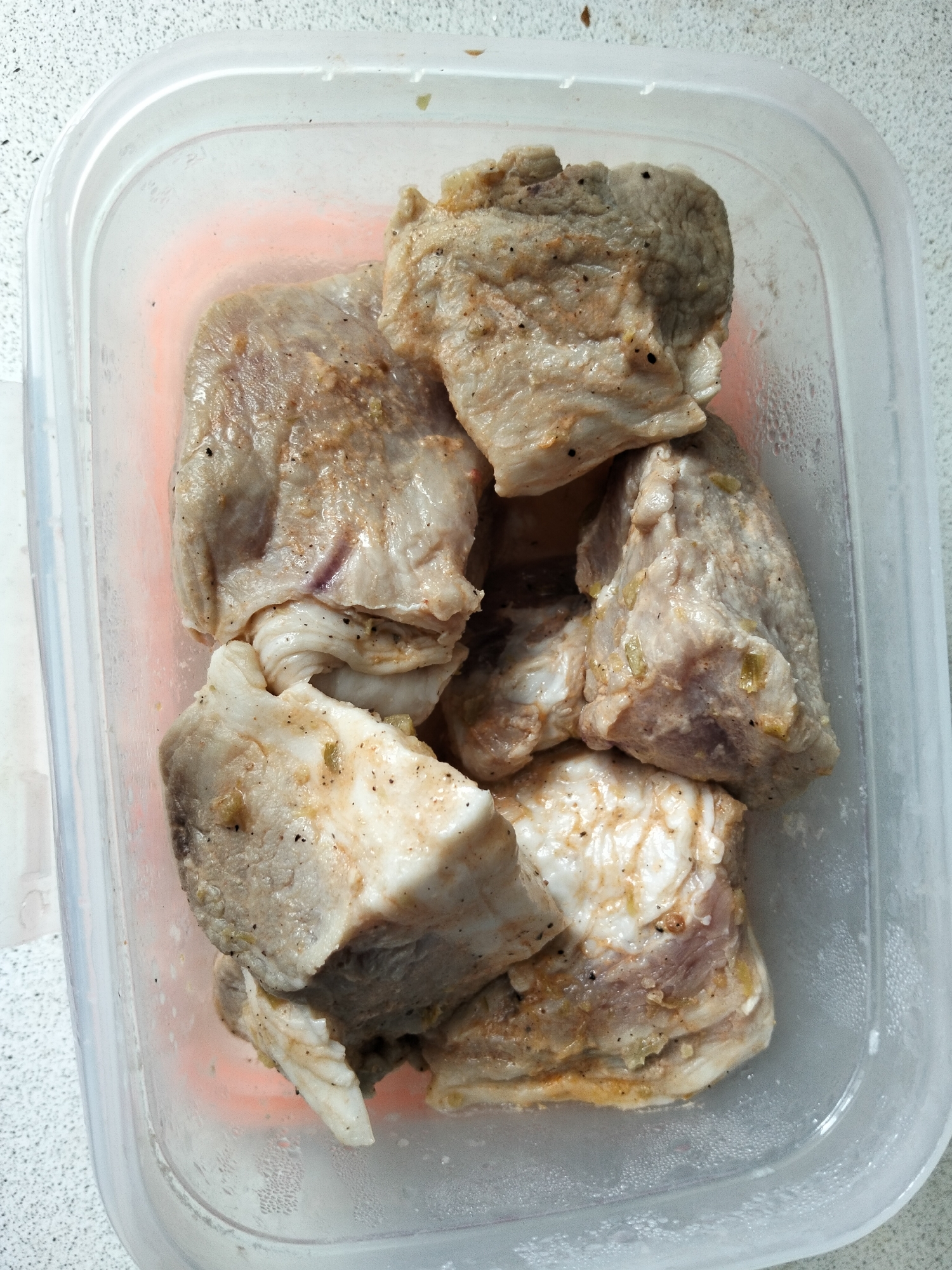 A clear plastic food storage container, filled with several pieces of raw, seasoned meat, sits on a speckled light-colored countertop. The meat, appearing to be pork due to its color and visible fat, is cut into bite-sized chunks and seasoned with black pepper and what appears to be dried herbs or spices. Some pieces show a reddish hue, possibly indicating marinades or spices. Condensation is visible on the inside walls of the plastic container, suggesting the meat is chilled. The countertop has a textured, speckled pattern. The background is out of focus, but suggests an indoor setting. The image is taken from a top-down perspective, capturing the contents of the container clearly. There are no people or animals present in the image. The overall scene is domestic and relates to food preparation.