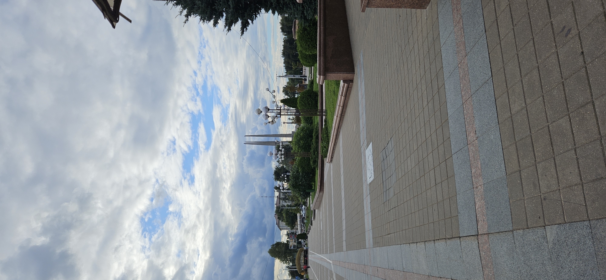 This is a vertical, medium-angle, outdoor, full-shot photograph of Victory Square in Vitsebsk, Belarus. The square is paved with square stone tiles, with a grey and pink granite border. On the left side of the frame, a walkway with a light-colored border runs parallel to a green grassy area with bushes and trees. There are a few people walking in the distance on the walkway. In the background, there are several buildings, trees, and two tall, pointed obelisks. The sky is filled with dramatic, dark grey clouds interspersed with patches of blue sky. Several ornate streetlights with multiple globe-shaped bulbs are visible in the middle ground. The overall scene suggests a public square in an urban environment, possibly with a monument commemorating a victory. The time of day appears to be daytime, likely late afternoon or early evening, given the cloudy sky.