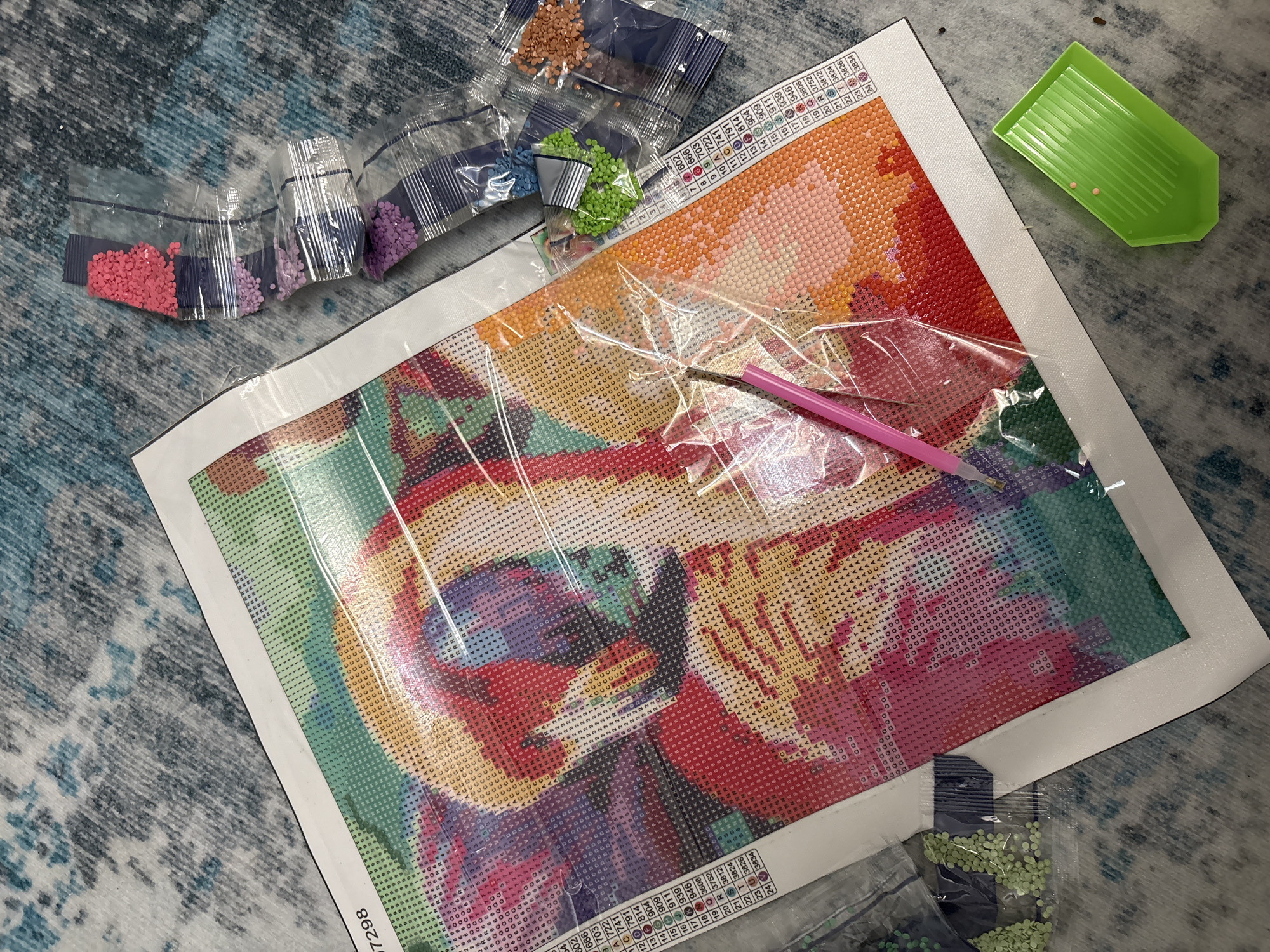An overhead shot captures a diamond painting kit laid out on a patterned blue and grey carpet or rug, likely indoors in Izhevsk, Russia. The central focus is a large canvas featuring a partially covered abstract or animal design, rendered in vibrant shades of red, orange, yellow, green, blue, and purple. The design is comprised of a grid of symbols and numbers, with a legend along the top and left edges indicating corresponding diamond colors.
Several clear plastic bags containing small, resin "diamonds" (also known as drills) are scattered around the canvas. These include distinct piles of pink, light purple, darker purple, light blue, orange, brown, and various shades of green diamonds. Some bags are open, revealing the contents.
A bright pink diamond painting pen is visible on the canvas, resting over a protective clear plastic film that covers the sticky surface of the design. A green, ridged plastic tray, commonly used to hold and align the diamonds, sits in the upper right corner of the frame, containing two small pink diamonds.
The canvas has a white border and part of a number, "97298," is visible along its bottom left edge. The legend on the canvas displays numbers from 1 to 24, each paired with a unique symbol (letters and numbers) and a four-digit DMC color code (e.g., 3604, 3824, 814, 911), allowing the crafter to match the diamonds to the correct spots on the canvas. The scene suggests an ongoing or recently started crafting activity, with tools and materials ready for use.