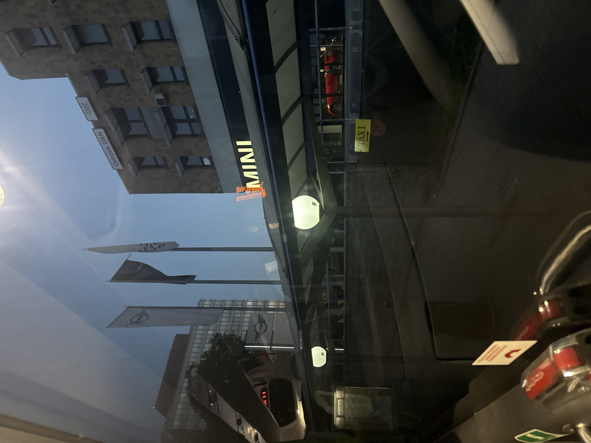 The image displays a street-level scene in Ostrava, Czechia, captured from the interior of a vehicle, likely a bus, evidenced by internal reflections of controls and a visible "Flixbus" logo reflection. The time of day appears to be either early morning or late evening due to the clear, pale blue sky and illuminated signs.
On the right side of the frame, a multi-story building with a light brown facade features numerous windows, some equipped with external blinds. Two white rectangular signs are mounted on its upper section: one reads "Ostrava. UAN" and the other "CSAD." Below these, a prominent, brightly illuminated white sign spells "MINI," indicating a MINI car dealership or showroom. An external air conditioning unit is visible on the building's facade.
Directly below the "MINI" sign, a covered walkway or shelter structure extends outwards, supported by poles. Two circular light fixtures, emitting a warm white light, are integrated into the underside of this overhead structure. Behind the glass frontage of the building, a red MINI car is visible on display within the showroom.
Further down and to the left of the car display, a yellow rectangular sign with a black border and text reads "TAXI" and includes an arrow pointing to the right, indicating a taxi stand or direction. A paved ground surface is visible beyond the lower walls.
To the left of the main building, three tall flagpoles are positioned. The flagpole closest to the building displays a white flag featuring the circular MINI logo. The central flagpole holds a dark grey or black flag, while the leftmost flagpole displays a white flag with an indistinct design.
The foreground is dominated by reflections on the vehicle's window, which partially obscure the external scene. These reflections include portions of the vehicle's interior, such as a seat back, red control buttons or levers, and a green emergency exit symbol. A small white sign with red, unreadable text is also reflected in the lower central part of the window. No individuals are visibly present in the scene.