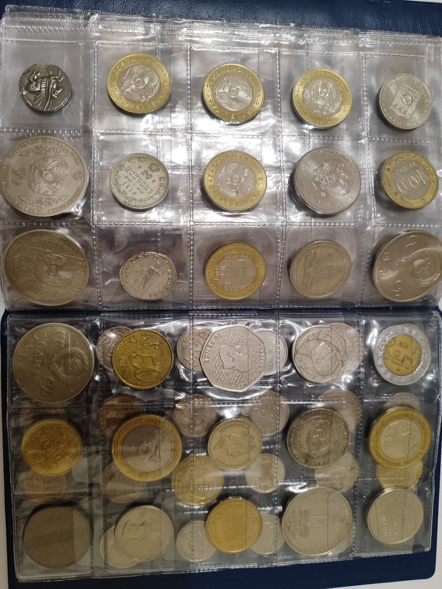 The image displays a collection of coins stored in plastic sleeves within a binder. The binder appears to be dark blue. The coins are of various sizes, colors, and designs, suggesting they are from different countries and time periods. Some coins are silver, while others are gold or a combination of both. The coins are arranged in rows and columns within the transparent plastic sleeves, allowing their obverse and reverse sides to be visible. There are no people or animals present in the image, nor any discernible environmental elements beyond the binder itself. The scene is static and focuses solely on the coin collection. There is no information present in the image to suggest a location in the city of Pavlodar, Kazakhstan.