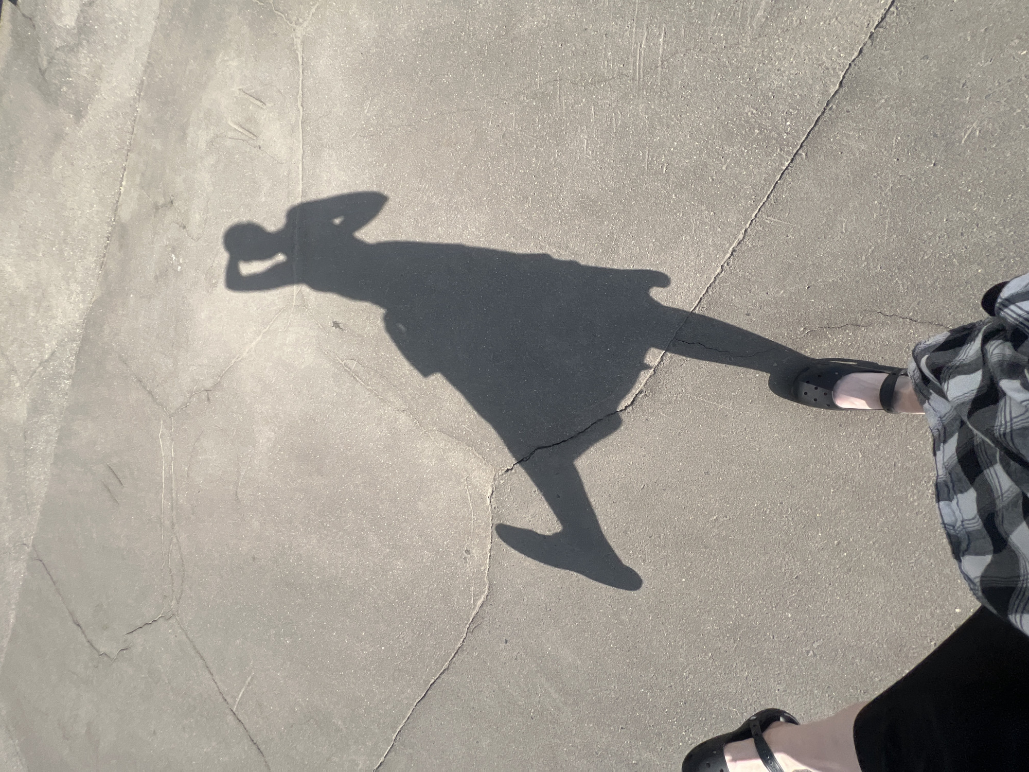 The image is a top-down view of a person's shadow cast on a concrete surface. The shadow appears to be of someone standing with their hand on their head. Part of the person's clothing is visible on the right side of the image. They are wearing a black and white checkered shirt and black leggings or pants. Two feet in sandals can also be seen. The background is a cracked, gray concrete surface. The location in the city of Khimki, Russia is not determined from the picture.