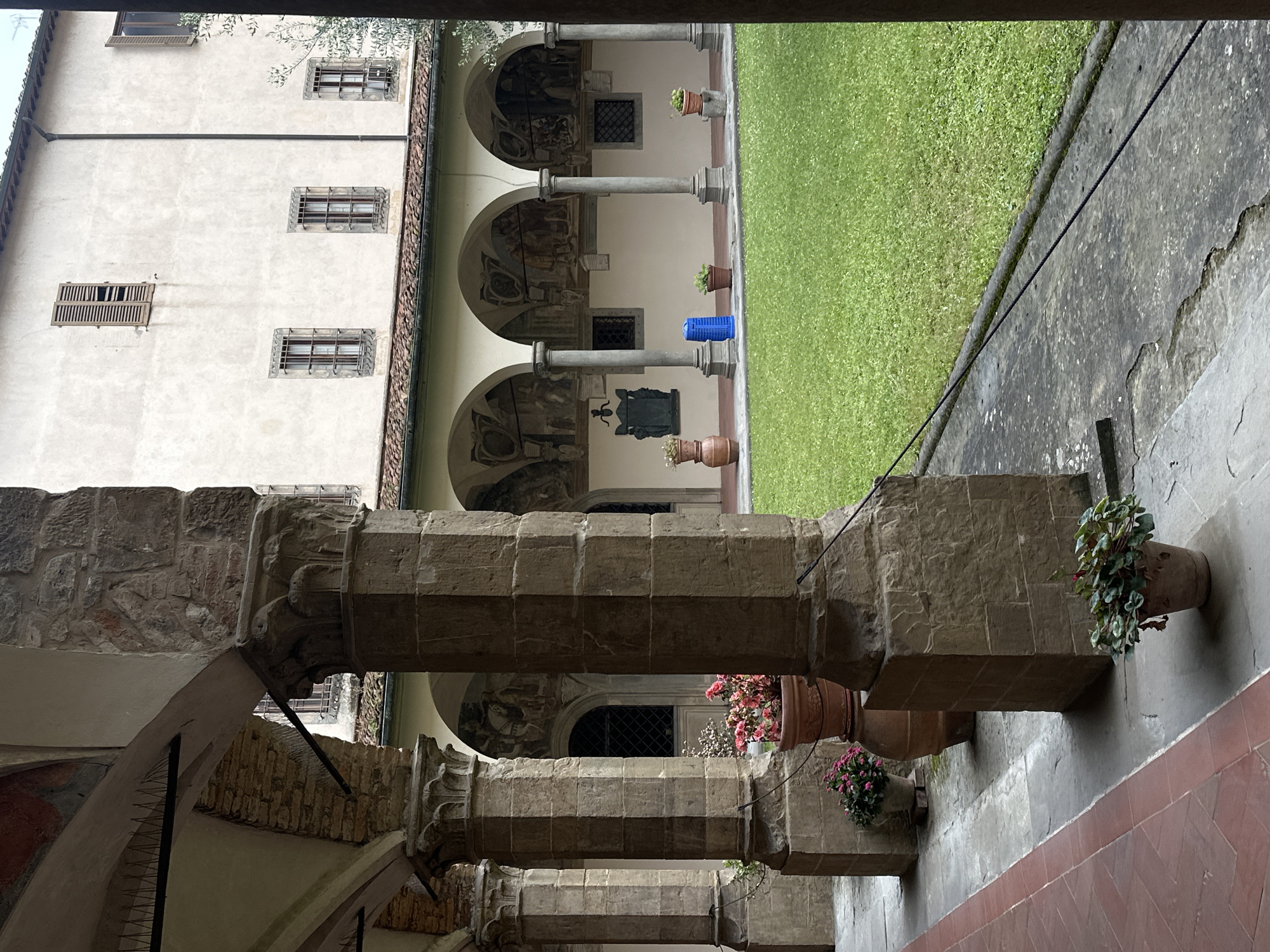 The image captures a serene view within a historic courtyard or cloister, characteristic of Florence, Italy. The perspective is from under a covered walkway, looking across a grassy central area towards an opposing building.
In the immediate foreground, a series of robust, octagonal stone columns support heavy stone arches, forming part of a shaded portico. Several terracotta pots, some containing vibrant pink and red flowering plants, and others green foliage, are placed on ledges alongside these columns. The floor of this walkway appears to be paved with reddish-brown tiles.
Beyond this foreground structure, a neatly manicured patch of green grass forms the central courtyard. On the far side of this grassy expanse, another architectural facade is visible. This building features a series of lighter-colored, round columns supporting arches, within which faded frescoes or murals are discernible. A dark statue or bust on a pedestal stands in one of these arched recesses. A bright blue cylindrical object, possibly a bin or barrel, rests on the grass near one of the columns.
Further in the background, a tall, light-colored stucco wall rises, pierced by multiple rectangular windows. Some windows are protected by decorative metal grilles, while one on a lower level features wooden shutters, partially open. A dark drainpipe runs vertically down this wall. Overhanging green foliage, possibly from a tree, is visible at the top of the frame.
The lighting is soft and diffused, suggesting an overcast day or a shaded environment. There are no people visible, and no specific activity is taking place, contributing to a calm and contemplative atmosphere. No discernible text is visible within the frame. The overall scene evokes a sense of historical grandeur and tranquility.