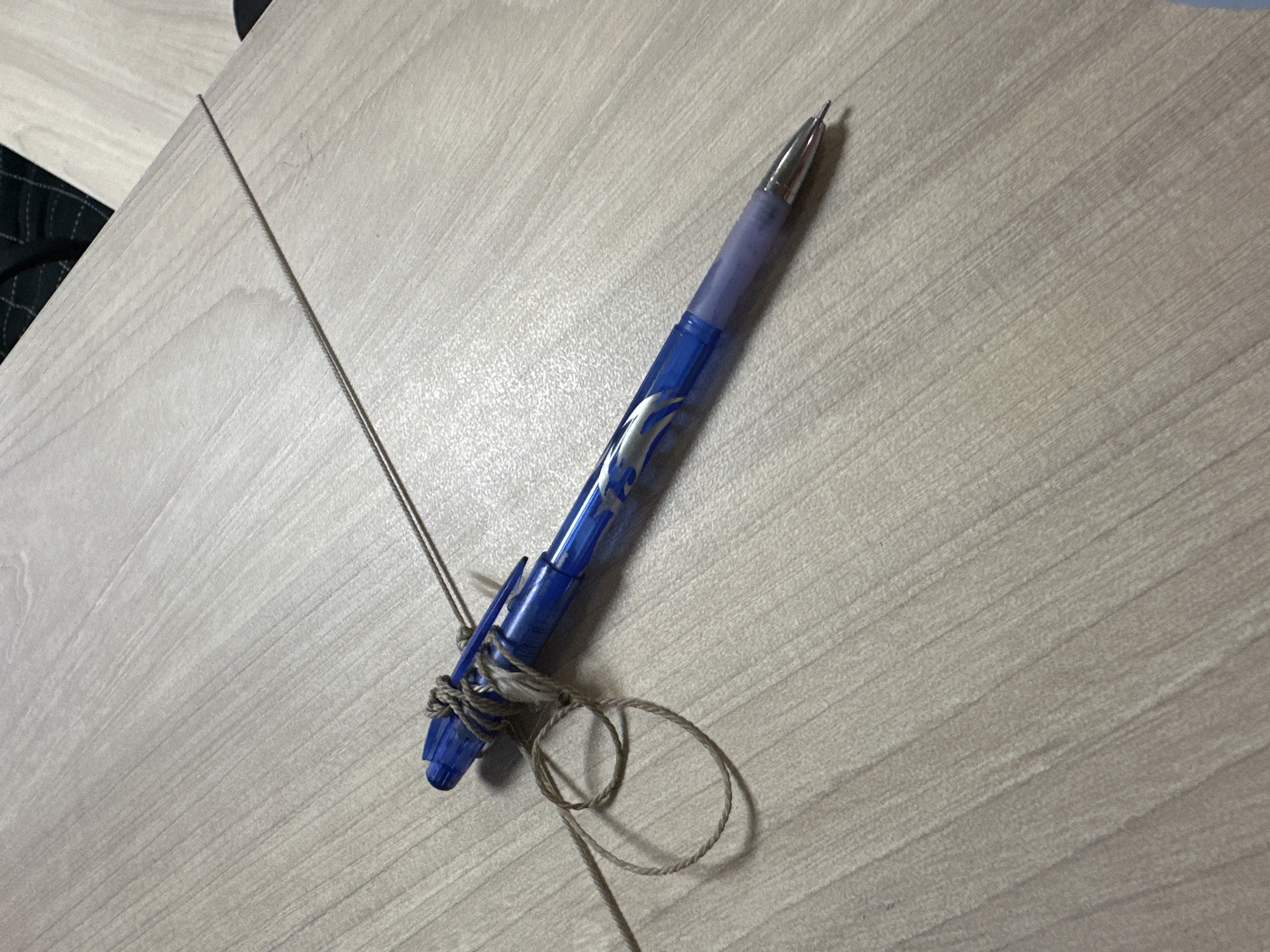 The image shows a blue pen with a silver tip, tied with twine. The pen is lying on a light-colored wooden surface, possibly a table, with visible wood grain. The twine is wrapped around the pen in several places and forms a loop on the surface to the left of the pen. The background is out of focus and shows a continuation of the wooden surface and possibly a dark fabric. There are no people or animals visible in the image. The scene appears to be indoors, with no indication of the city of Hora, Ukraine.