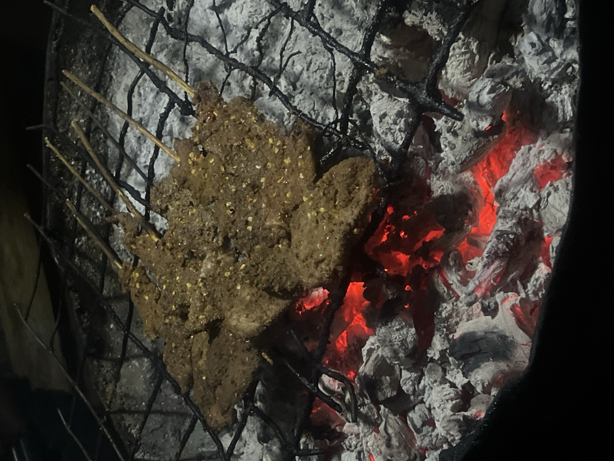 The image depicts a close-up view of meat skewers being grilled over hot coals. The skewers, each containing several pieces of seasoned meat, are arranged on a metal grill rack. The meat appears to be seasoned with a mixture of spices, visible as small flecks on its surface. Beneath the grill, glowing red coals provide the heat for cooking. The surrounding environment is dark, suggesting an outdoor setting, possibly at night. The scene is characteristic of a food stall or an outdoor cooking event in a location such as Durimi, Nigeria, where grilled meats are a popular culinary offering.