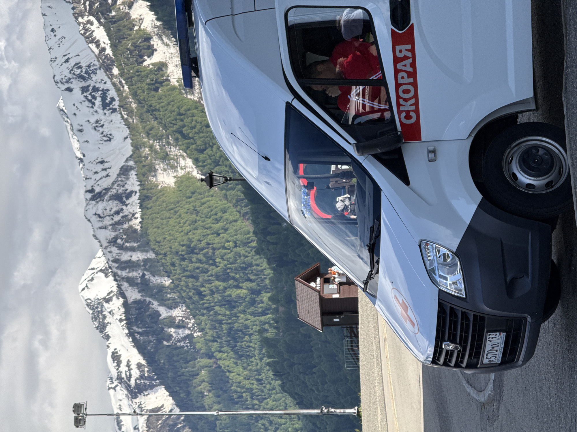 A white ambulance van is prominently featured, parked on what appears to be an asphalt surface in a mountainous region. The vehicle is oriented with its front facing slightly towards the right and the snowy mountains in the background.
The ambulance is white with a red stripe along its side, on which the Russian word "СКОРАЯ" (SKORAYA), meaning "Emergency" or "Ambulance," is clearly visible in white letters. A red cross symbol is present on the front hood. The front license plate reads "К570МХ 193 RUS". Inside the driver's cabin, a person wearing a red jacket is seated. A blue flashing light is mounted on the roof of the vehicle.
In the background, majestic snow-capped mountains rise under a partly cloudy sky, with their lower slopes covered by dense green coniferous forests. A small, dark-roofed wooden building is nestled among the trees on a slope, suggesting some form of mountain infrastructure. A tall lamppost or light pole stands on the left side of the frame.
The scene takes place during the daytime, with clear visibility. The combination of snow-covered peaks and green forests indicates a high-altitude location, likely in late spring or early summer. This setting is consistent with the provided context of Estosadok, Russia, a mountain resort area in the Caucasus. The image captures a static moment, with the ambulance seemingly stationed amidst the scenic, rugged landscape.