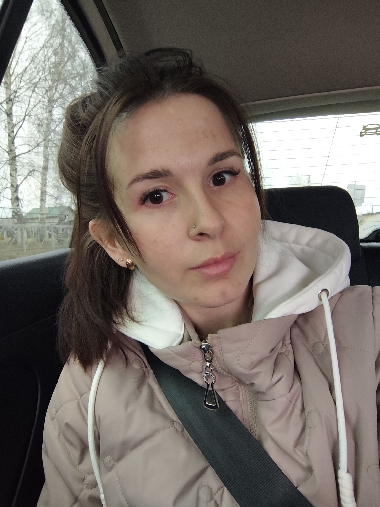 A woman is in the back seat of a car. She is wearing a seatbelt and a light pink quilted jacket with a white hood. She has brown hair, pulled back, and a nose piercing. Trees and buildings are visible through the car window, which allows one to determine the location is Фоки, Russia.