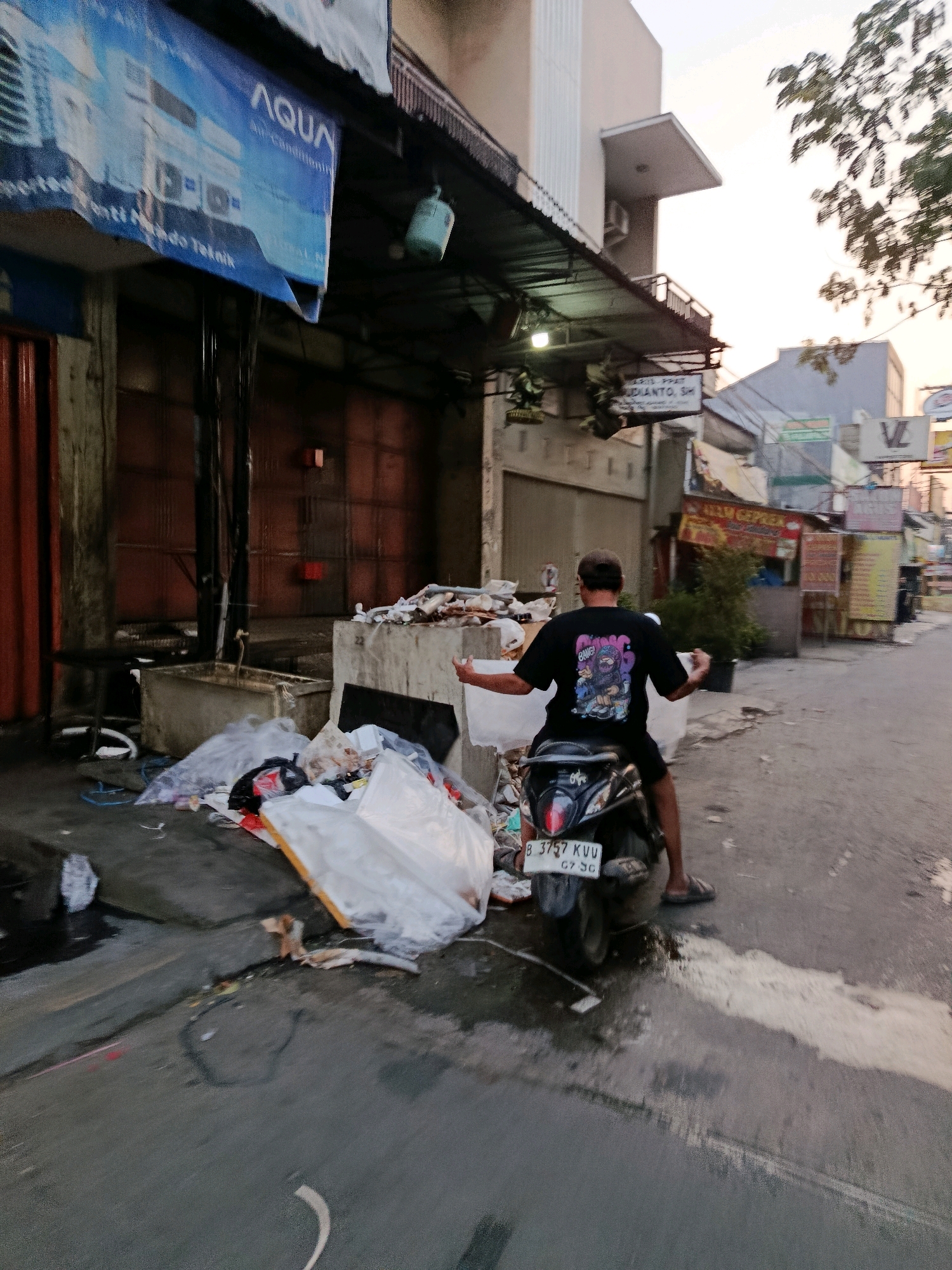 In Bekasi, Indonesia, a scene unfolds on a street cluttered with debris. A man, wearing a black t-shirt with a graphic print and dark shorts, is seated on a black motorcycle. He is facing away from the camera, with his arms outstretched to his sides. The motorcycle is parked on a paved surface, partially obscured by a large pile of garbage bags and assorted trash. This debris is piled high next to a concrete structure, suggesting a neglected area. In the background, buildings line the street, some displaying signs with text in Indonesian, indicating a commercial or residential area. The sky above is overcast, casting a soft light on the surroundings. The overall impression is one of urban disarray, with a focus on the accumulation of waste in a public space.