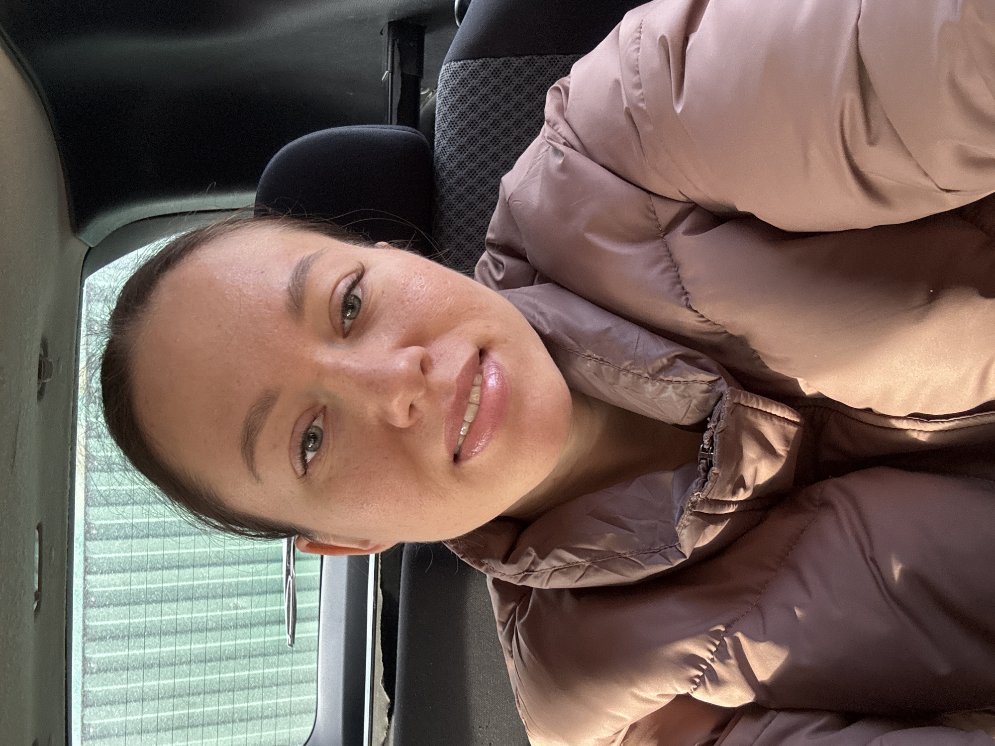 An adult female is shown in a selfie taken from the rear passenger compartment of a vehicle in the city of Ферма, Russia. She wears a puffy, salmon-pink winter jacket. Her brown hair is pulled back, her light-colored eyes are open, and her lips are slightly parted, revealing her upper teeth. To her right, a black vehicle headrest and a black seat with a dark grey patterned insert are visible. The vehicle's rear window shows a blurred exterior background of a vertically striped green structure. The car's ceiling and an overhead grab handle are present at the top of the frame.