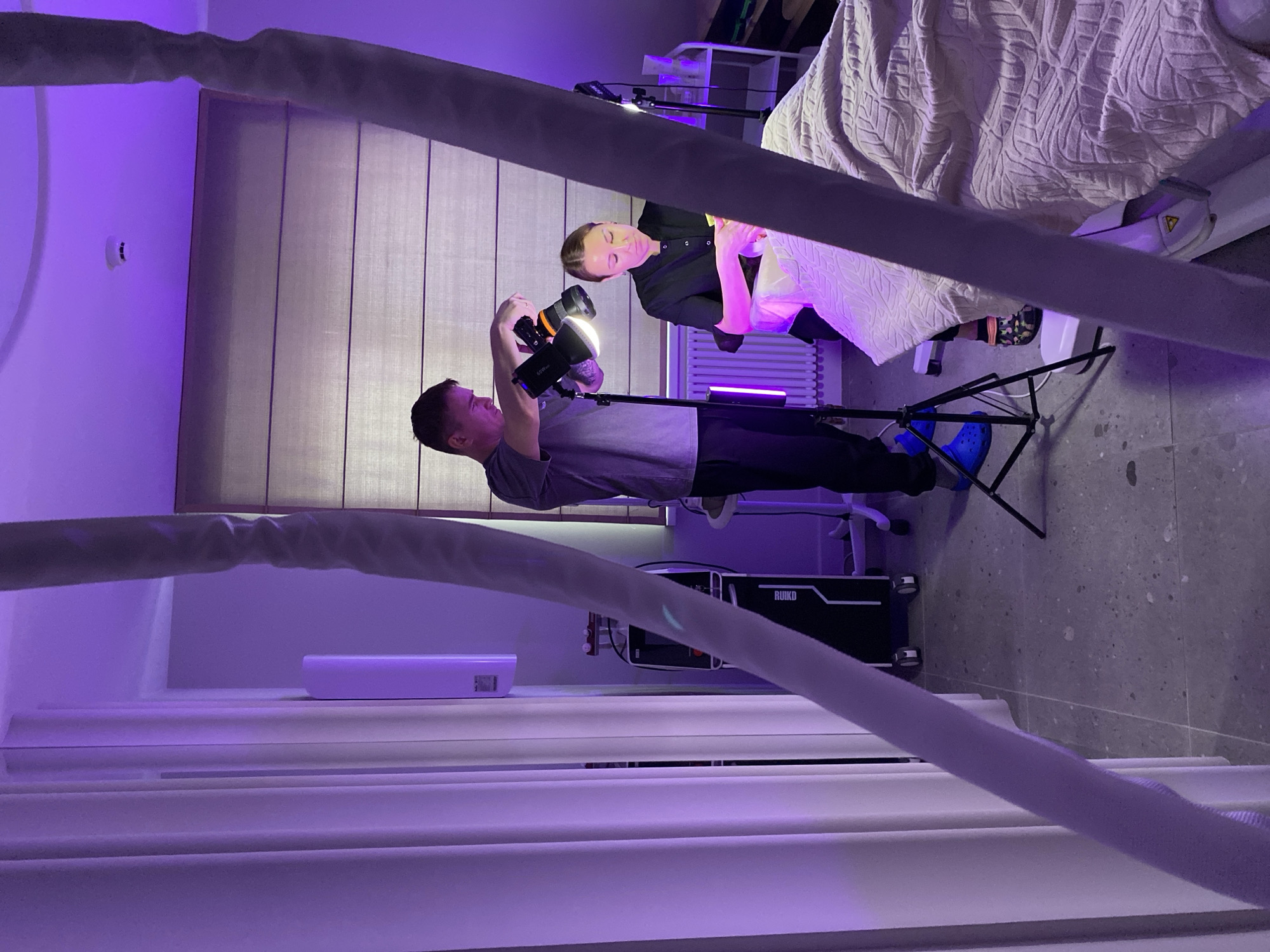 Two people are in a room with purple lighting, likely a studio. A man in a gray shirt, black pants, and blue shoes is holding a camera with a flash attachment, taking a photograph of a woman wearing a black top. The woman is lying on a bed covered with a patterned blanket. There is lighting equipment, including a light tube and tripods, visible in the background. The location in Izhevsk, Russia, cannot be determined from the image.