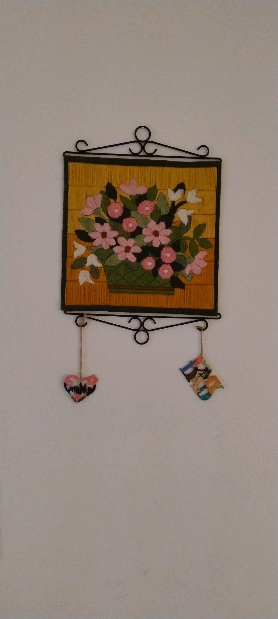 A framed needlepoint tapestry depicting a floral arrangement hangs on a plain, off-white wall. The tapestry, with a yellow background and a green basket overflowing with pink and white flowers, is enclosed in a dark border and suspended by a black metal scrollwork frame. Below the tapestry, two small, decorative items hang from the frame by strings. On the left is a heart-shaped ornament with a floral pattern. On the right is a cluster of colorful, three-dimensional letters that appear to spell out "DAD". The scene is indoors, and there is no visible indication of the specific location in Ishøj, Denmark, or any people present.