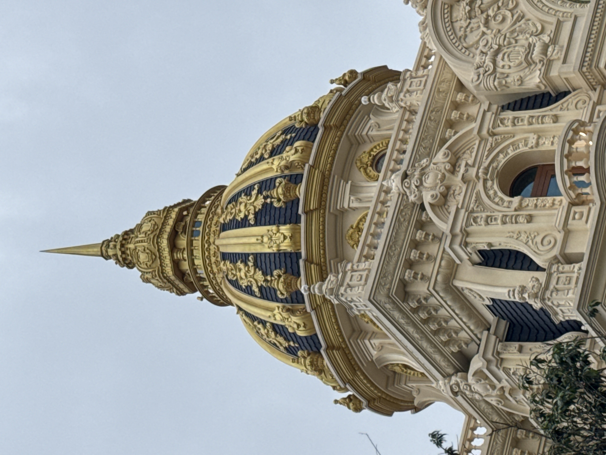 The image shows the ornate golden dome and upper facade of a building, likely a historical or religious structure, against a pale, overcast sky. The dome is adorned with intricate gold filigree over dark blue tiles, topped by a tall, pointed spire. The facade below the dome features detailed cream-colored stonework, including decorative carvings, arched windows, and balconies. Green foliage from trees is visible on the left side of the frame, suggesting an outdoor setting.
This building is identified as the Dinh 1 Bao Dai, also known as the Bao Dai's Summer Palace, located in Da Lat, Vietnam. Da Lat is a city in the Central Highlands of Vietnam, known for its French colonial architecture and cooler climate. The Dinh 1 is one of the palaces formerly owned by Emperor Bao Dai, the last monarch of Vietnam.
