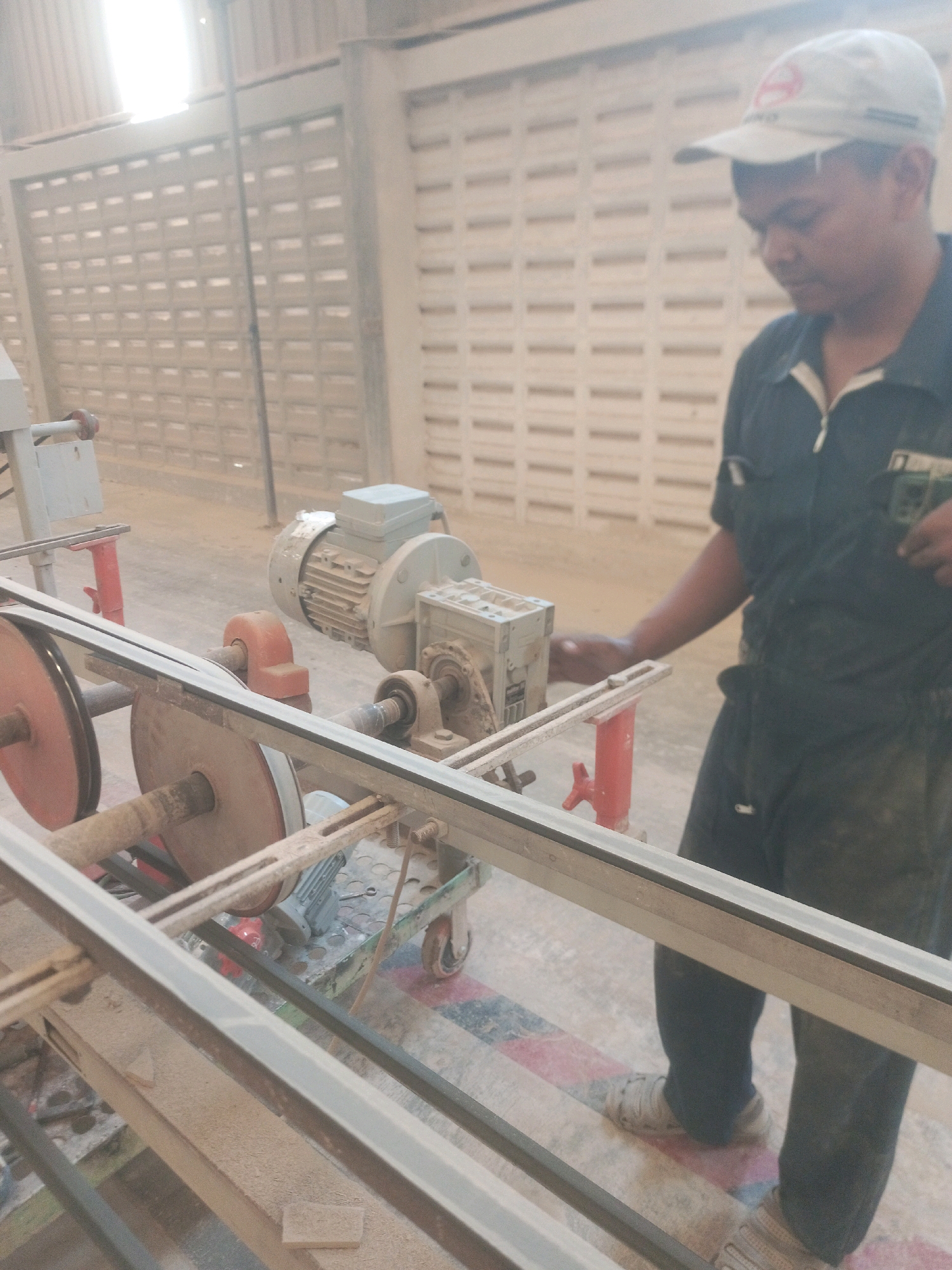 In Lebaniwaras, Indonesia, a man wearing a light-colored cap and dark work clothes stands near a mechanical saw in an industrial setting. He is looking down, seemingly focused on the machine. The saw has a silver motor, a red support structure, and two large pulleys with a belt running between them. The environment appears dusty, with a patterned floor and a wall made of rectangular blocks in the background. The lighting suggests an indoor or covered area with natural light coming from the left.