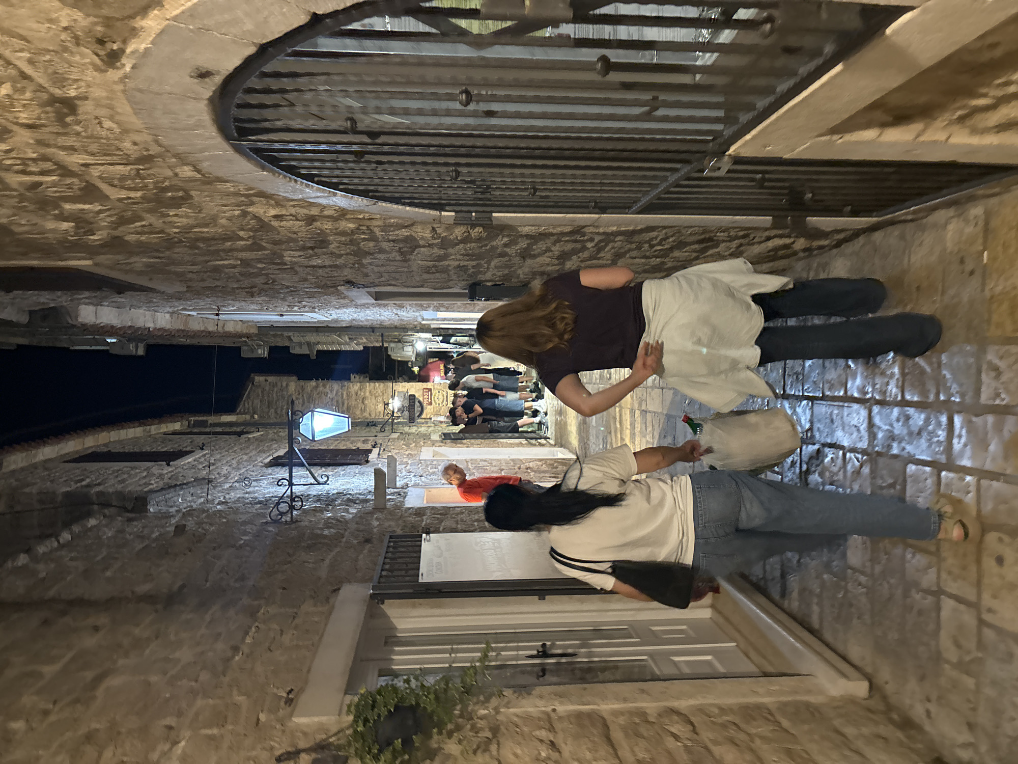 The image shows a narrow pedestrian street in the old town of Budva, Montenegro, at night. The street is paved with stone. Buildings line both sides of the street. Two women are walking along the street. One woman has long brown hair, wearing a dark-colored top, jeans, and a white skirt. She has her arm outstretched. The second woman has long dark hair, wearing a white and black striped t-shirt, jeans, and carrying a plastic shopping bag with a green bottle inside. A group of people is further down the street. A streetlight is mounted on a building. A metal grate spans the street overhead. A closed white door is on the left. A green plant grows next to the door.