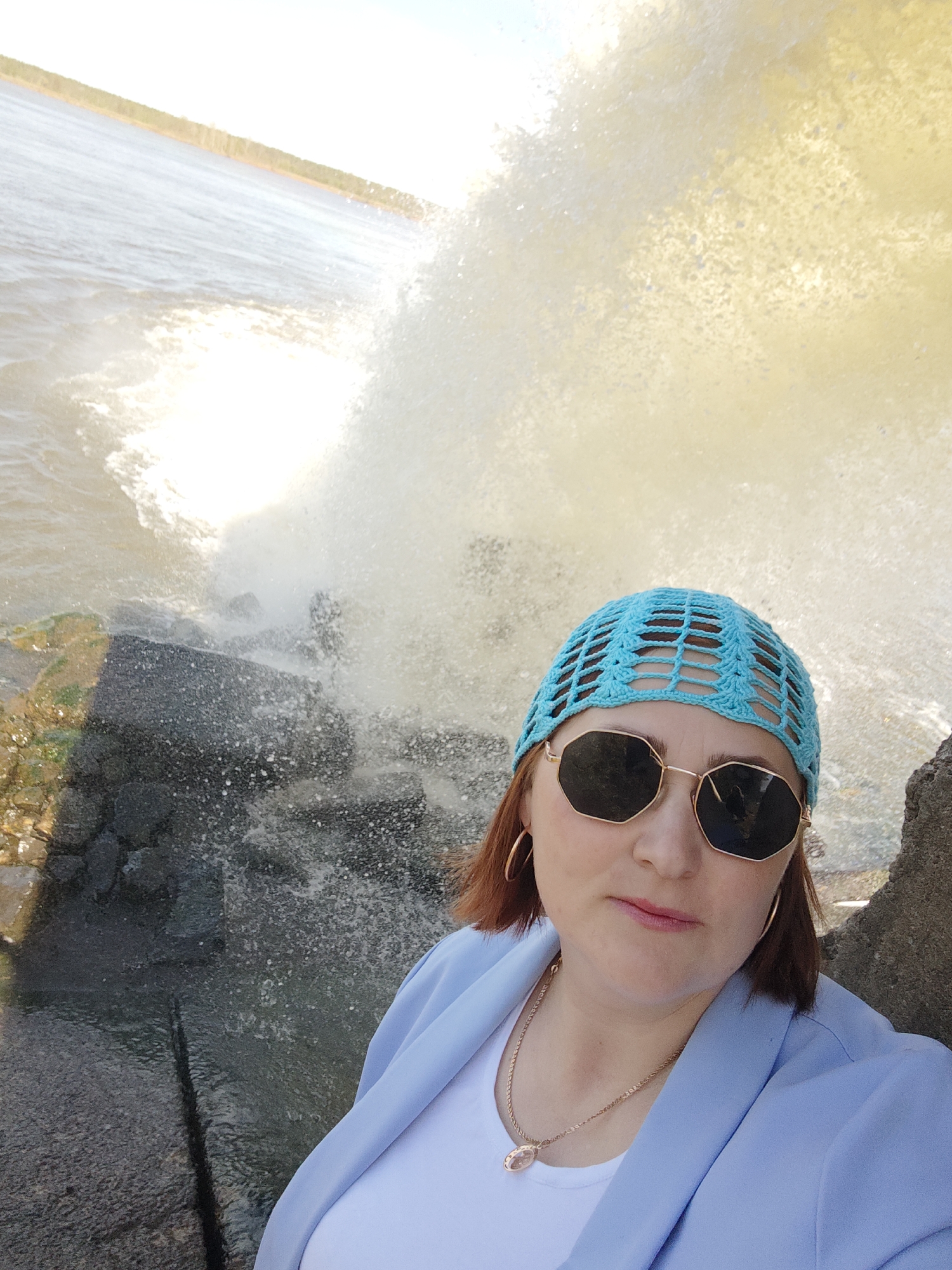 A selfie shows a woman with red hair, wearing a light blue crocheted hat, gold octagonal sunglasses, gold hoop earrings, a gold necklace with a pendant, a white t-shirt, and a light blue blazer. She is standing outdoors near a body of water where waves are crashing against rocks. The location is likely Chernushka, Russia, based on the assumption that the request to provide that location indicates the photo was taken there. The light suggests it is daytime.