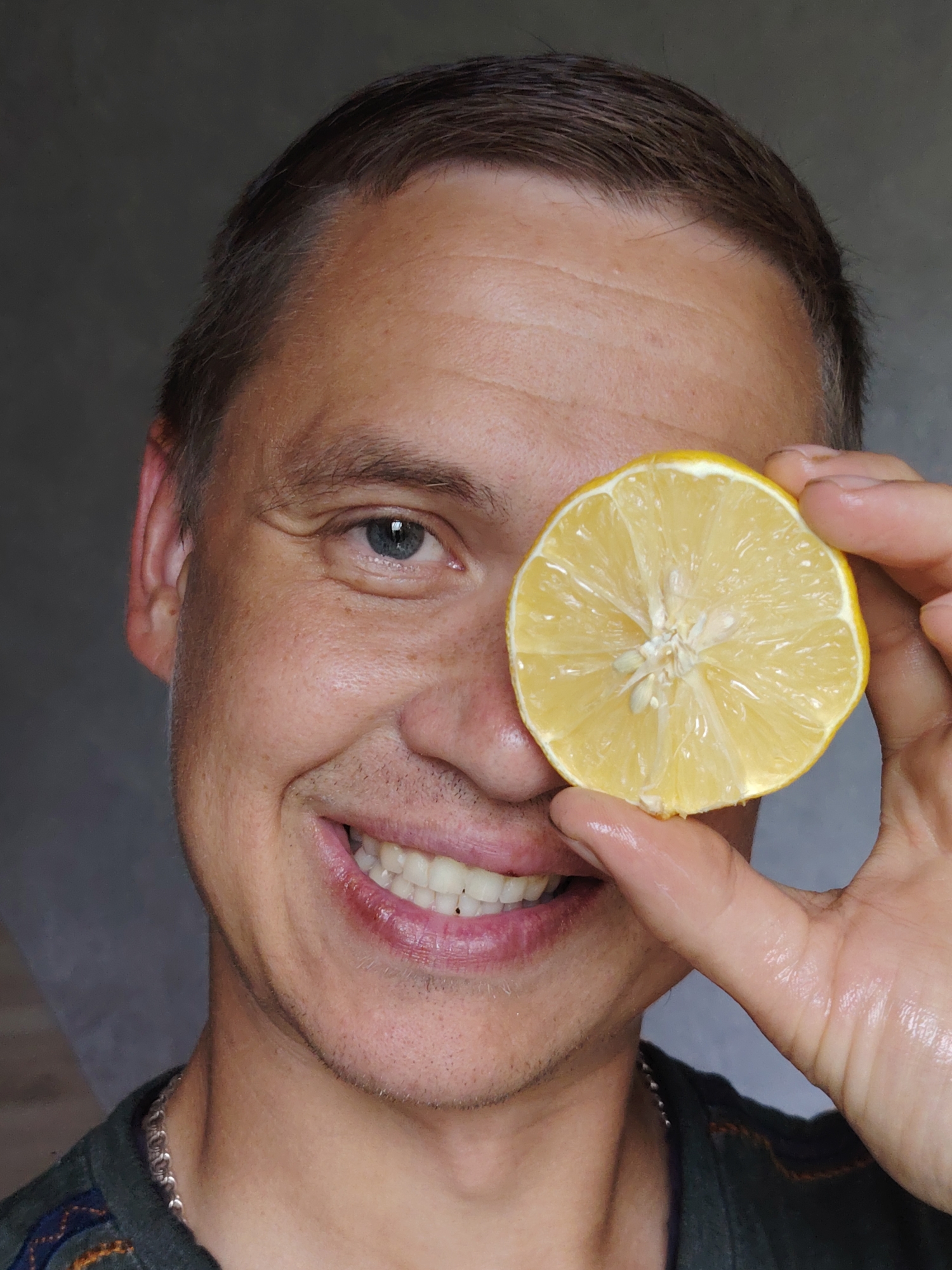A close-up, waist-up shot shows a man in his 30s holding a slice of lemon over his right eye. He is smiling broadly, revealing white teeth. The man has short, dark hair and blue eyes. He is wearing a dark green shirt with a patterned neckline and a silver chain necklace. The background is a plain, dark grey wall. The lighting suggests an indoor setting, possibly a home or studio, with no discernible indicators of the city of Chaikovsky, Russia.
