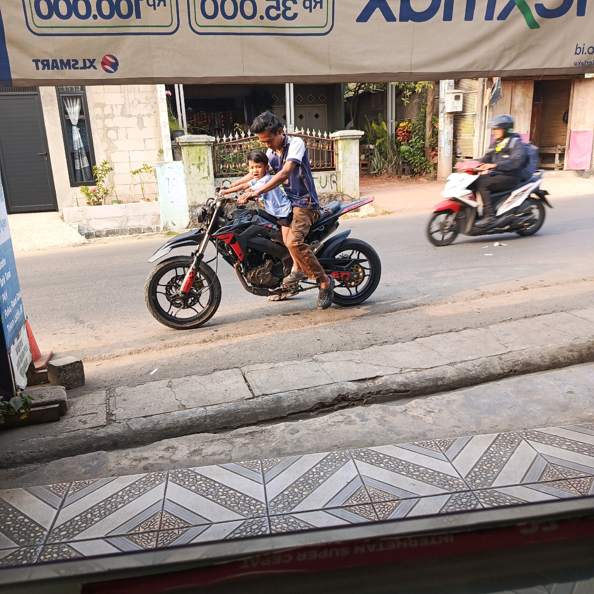 In Karawang, Indonesia, two individuals are riding a black sport motorcycle on a paved road. The driver, a young man, wears a dark t-shirt with blue and white accents and brown pants. He is looking forward while operating the motorcycle. Behind him, a younger boy, wearing a light blue t-shirt, holds onto the driver's shoulders. The motorcycle has red accents and a visible "DUNLOP" tire. In the background, another motorcycle, a white and red scooter, is being ridden by a person wearing a dark jacket and helmet, moving in the same direction. The scene is bordered by a sidewalk with patterned tiles and a raised concrete curb. Behind the road, buildings with light-colored brick walls and dark framed windows are visible, along with some greenery and fences. A large banner with blue and green lettering hangs above the scene. The lighting suggests it is daytime.