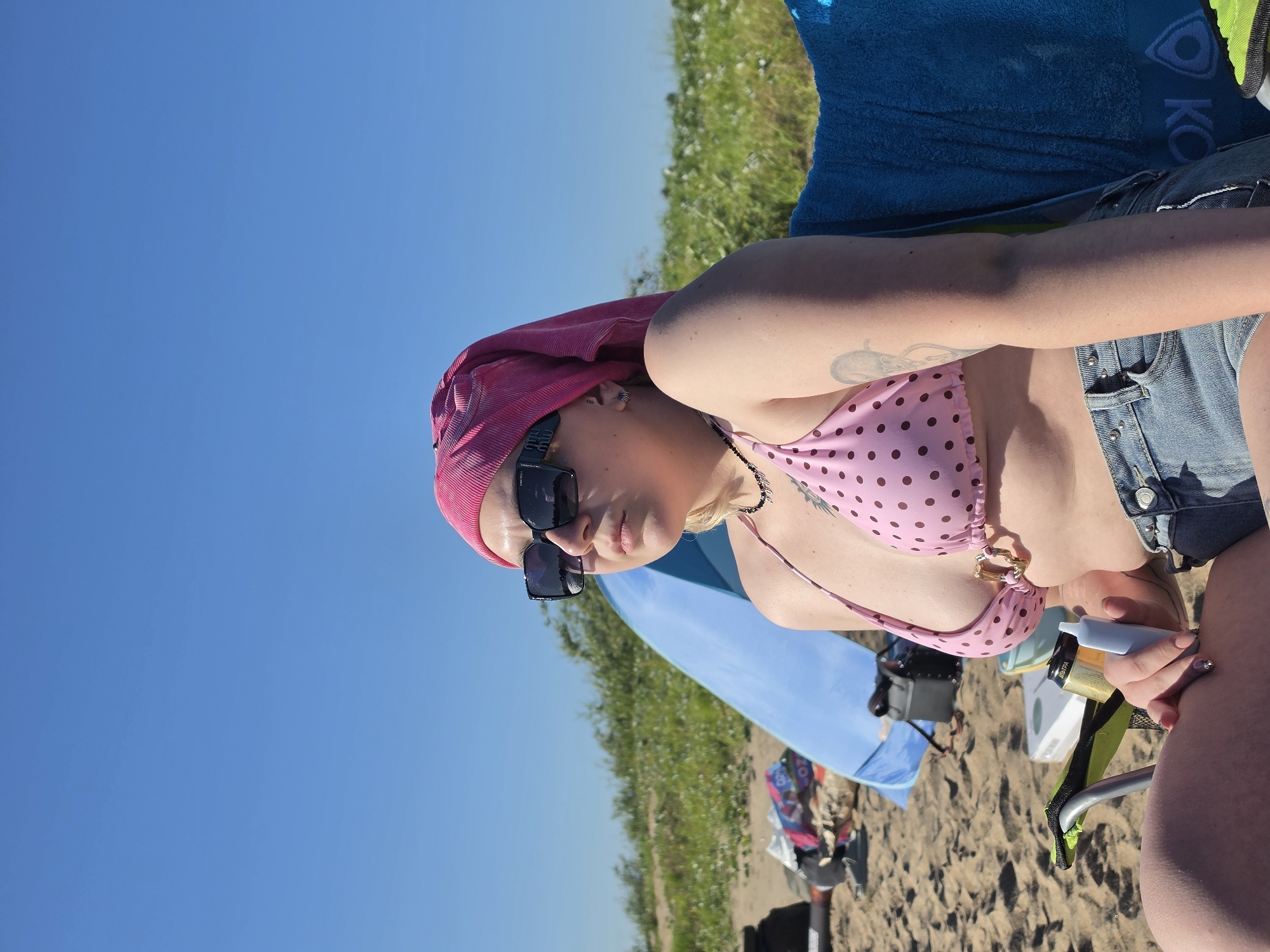 A young woman with blonde hair, wearing a pink polka dot bikini top, denim shorts, and a pink headscarf, sits on a beach chair under a clear blue sky. She is wearing large black sunglasses. Behind her, a blue beach tent is set up on the sandy shore, with some belongings scattered around. The background features a dune covered in green grass. The woman is holding a light blue object in her hand. The scene appears to be a sunny day at the beach, possibly in the city of Nizhny Novgorod, Russia, though this cannot be definitively confirmed from the visual information alone.