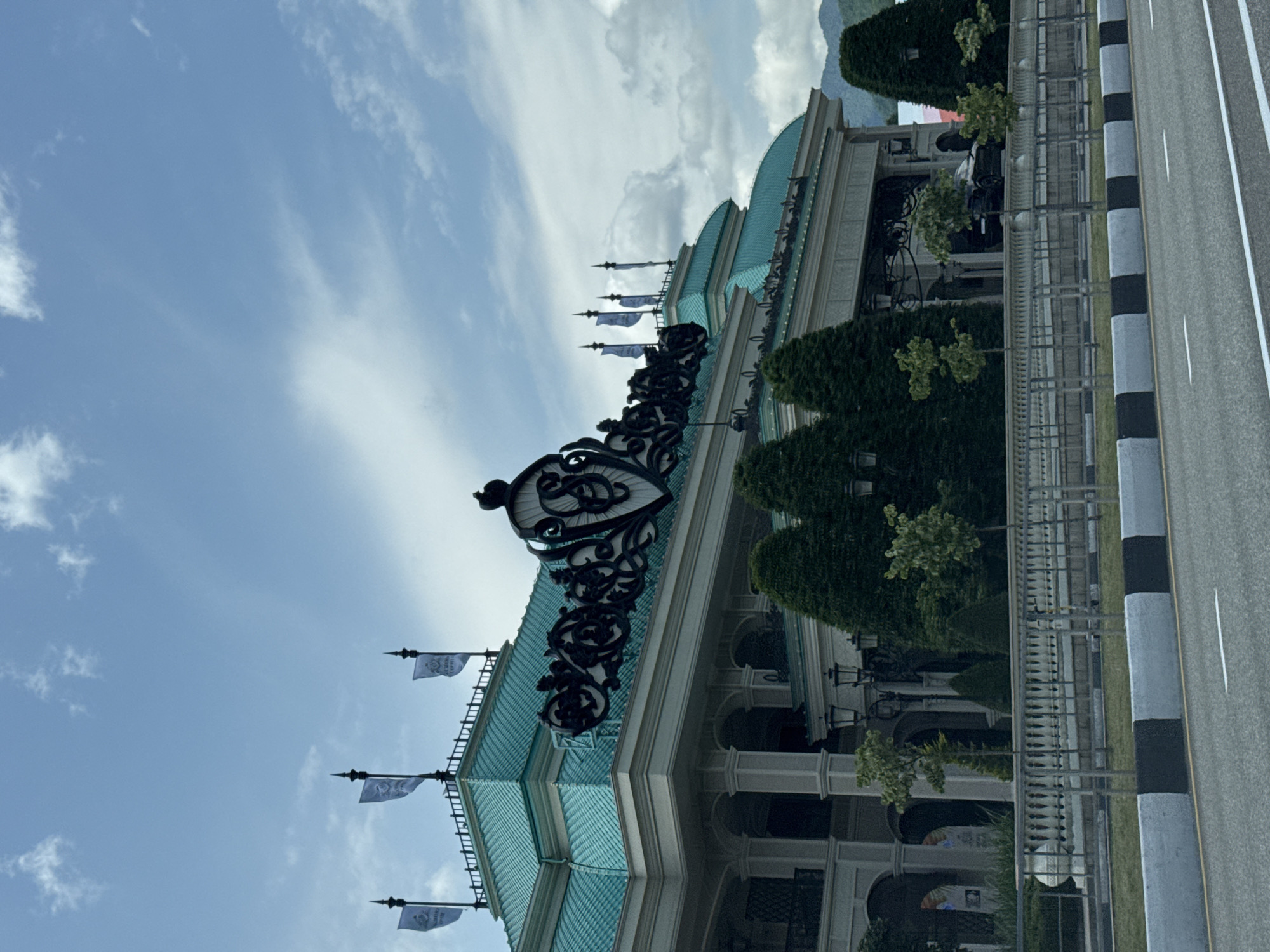 The image captures the facade of a grand building with a turquoise tiled roof and intricate black decorative elements. Several flags with a crest are visible on the rooftop. The building features large arched entrances, columns, and manicured trees in front of it. A white railing separates the property from a road in the foreground, which has black and white concrete barriers. The sky is partly cloudy, and mountains are visible in the background. The scene appears to be in Estosadok, Russia, likely a resort or hotel. There are no people or explicit actions depicted in the image.