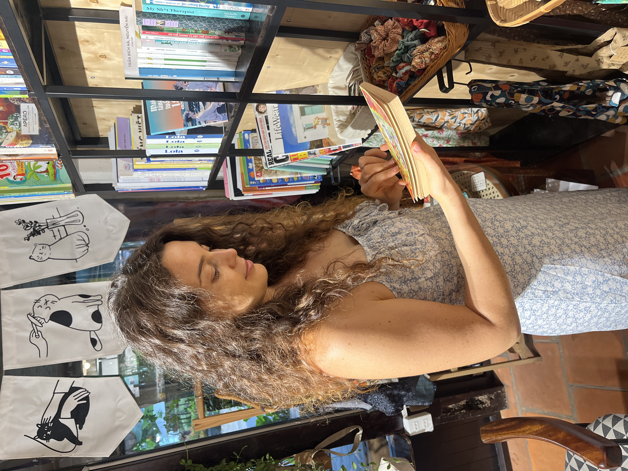 A medium shot captures a woman with curly brown hair wearing a light blue floral print dress as she browses a shelf of books in what appears to be a shop. Her gaze is directed downwards at a book she holds open in her hands. Behind her, a black metal shelf unit is filled with various books and magazines. Below the shelf, a wicker basket overflows with colorful fabric scrunchies. The background features a window with greenery visible outside, suggesting an outdoor element to the location, and decorative banners with black and white cat illustrations hanging above the bookshelf. The overall lighting is warm and natural, indicating it is daytime. The environment suggests a retail space focused on books and accessories.