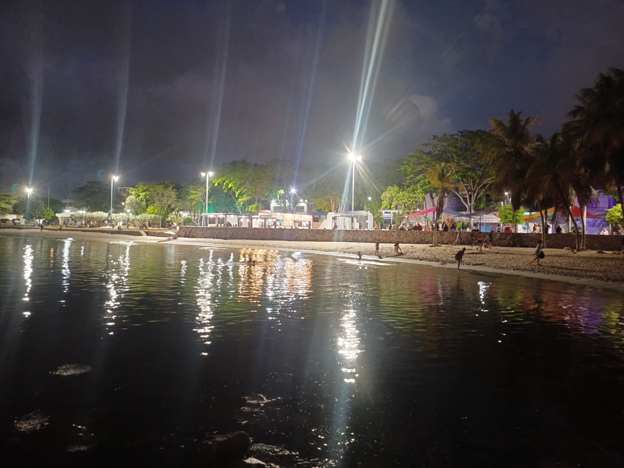 A nighttime scene depicts the beach area of Juan Griego, Venezuela. The foreground is dominated by dark water with reflections of lights from the shore. Several scattered rocks are visible in the water. The beach itself is sandy and illuminated by artificial lights, casting bright streaks across the scene. Along the shore, there are structures that appear to be stalls or open-air establishments, with people gathered around them. Palm trees and other foliage line the background, with more buildings and lights further back. A few individuals are visible on the beach and wading in the shallow water. The sky is dark with some cloud cover, and the overall impression is of a lively evening gathering.