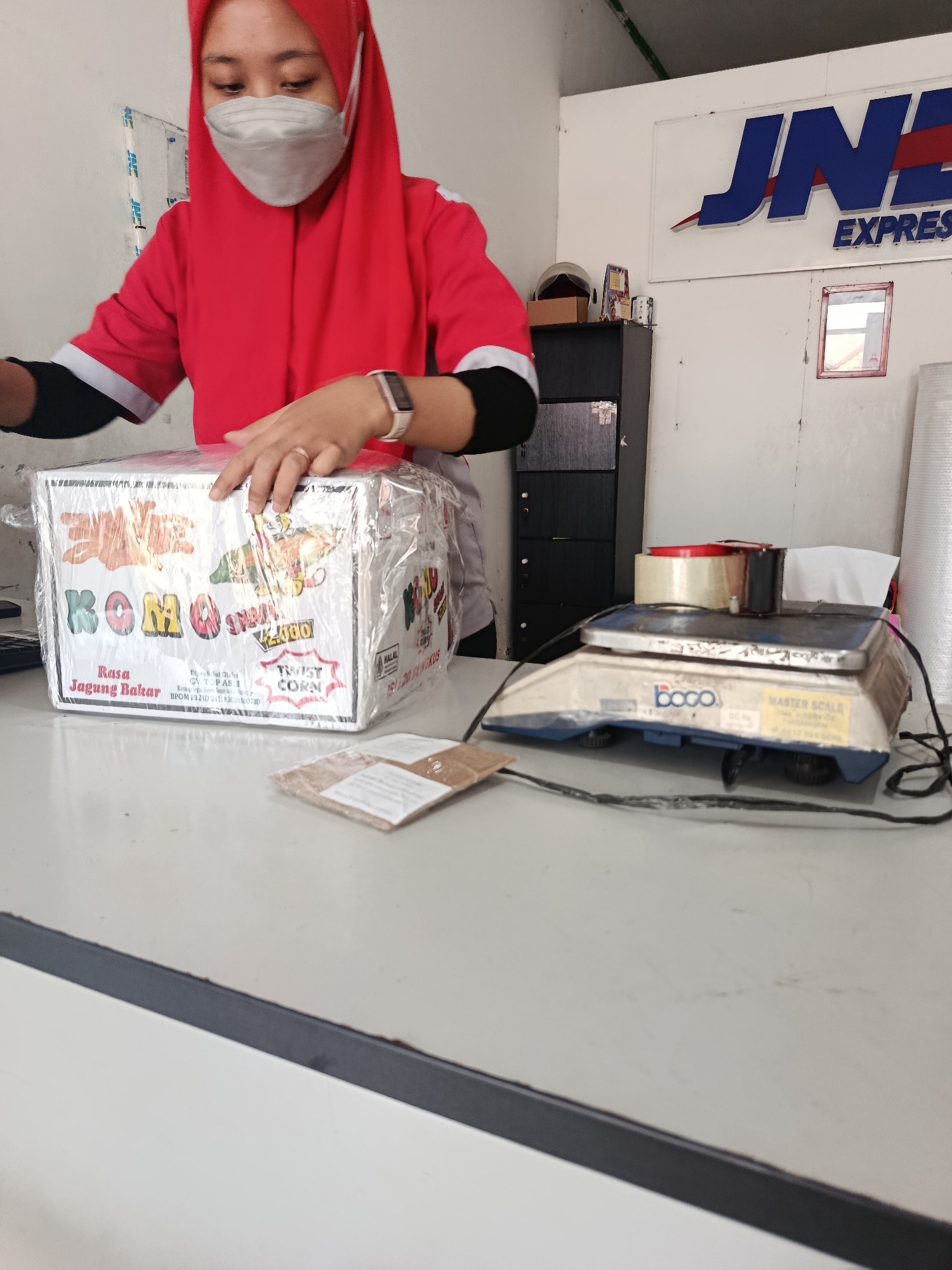 In Bantarsari, Indonesia, a woman wearing a red hijab and a red uniform with black trim is preparing a package. She has a face mask on and is using her hands to wrap a box in plastic. The box is decorated with colorful images and text, including the brand "Komo" and "Twist Corn." The woman has a ring on her left hand and a smartwatch on her right wrist.
The background shows a white wall with a large blue and red "JNE EXPRESS" logo. To the left, a dark cabinet is visible, with a helmet and some papers on top. To the right, a blue and white digital scale, labeled "Boco MASTER SCALE," is present, with rolls of tape on top. The scale is connected to a wire. In the foreground, a white table is shown, with a small brown package placed on it. The overall scene suggests a logistics or shipping environment.