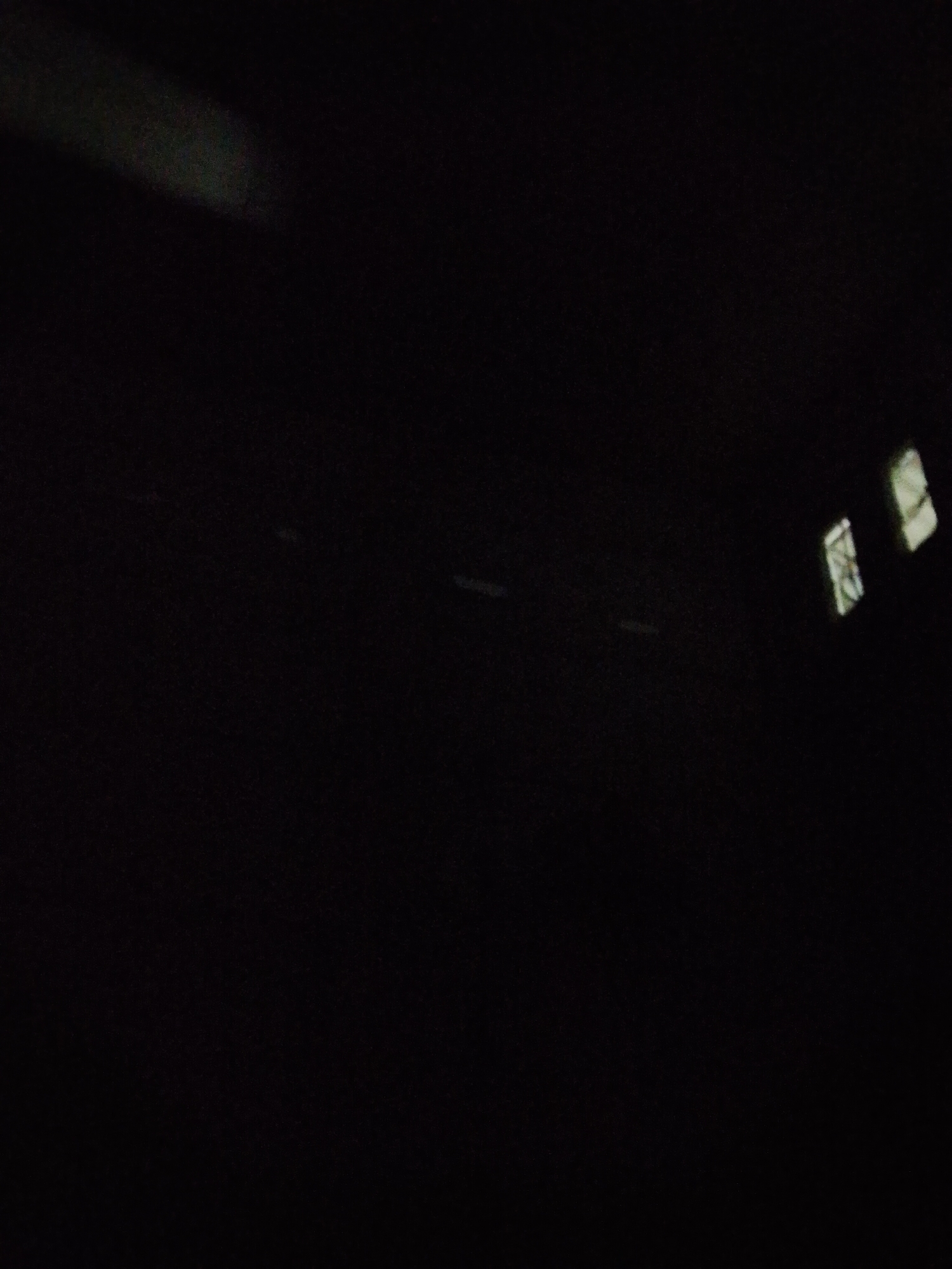 The image depicts a dark indoor space. Faint light emanates from what appears to be two windows on the right side of the frame. The windows appear obscured or partially covered, casting indistinct patterns of light. In the lower-middle part of the image, small amounts of light are visible, indicating a possible floor. The location is Bukit Ketok, Indonesia. No people, specific objects, actions, or interactions are visible.