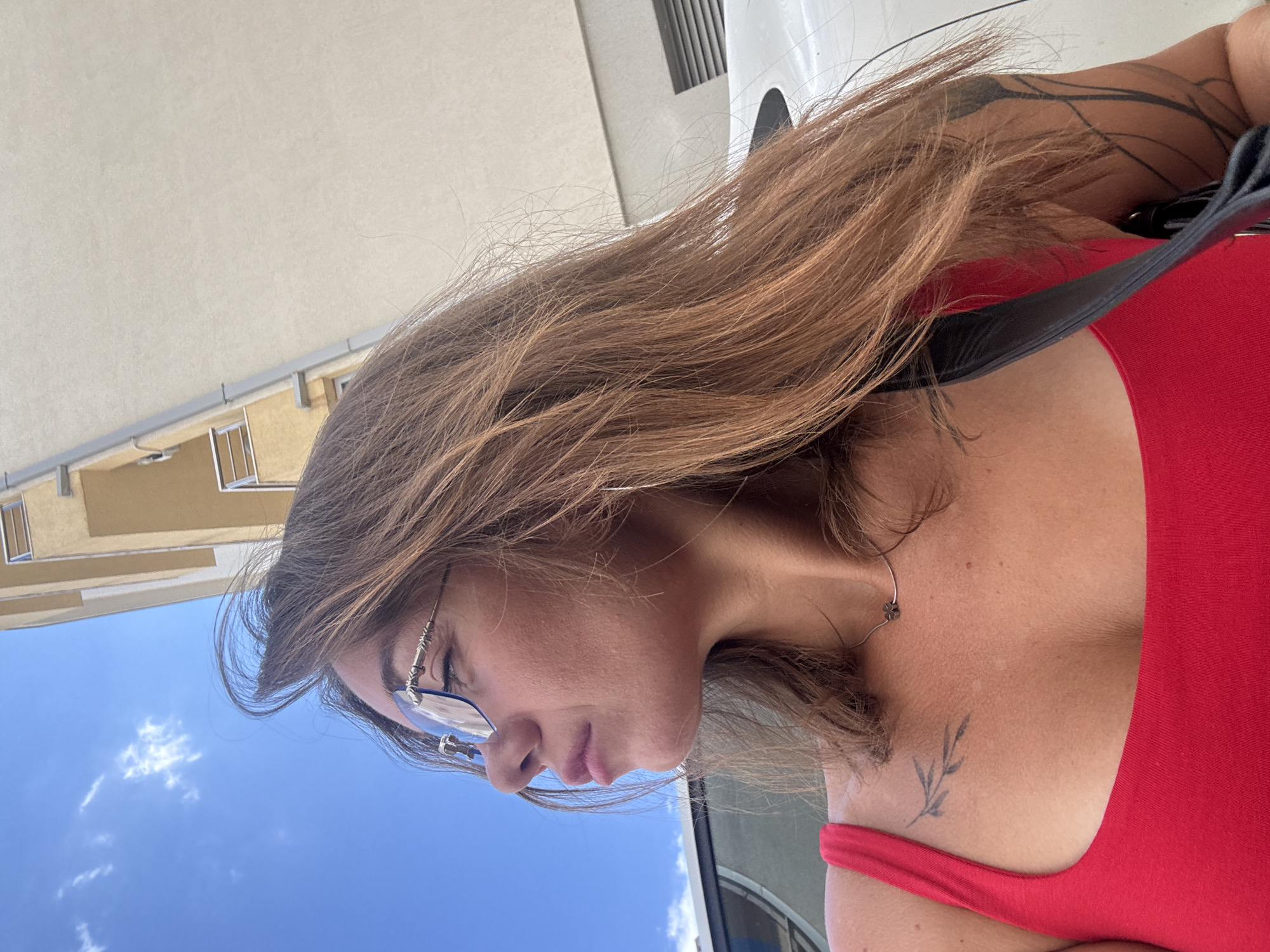 A medium shot captures a woman from the chest up, looking to her left. She has shoulder-length, light brown hair, bangs, and is wearing rimless glasses. Her skin tone is fair with freckles. She is wearing a bright red tank top, and a thin silver necklace with a four-leaf clover pendant. A small branch tattoo is visible on her left collarbone. In the background, a pale yellow building with balconies and a clear blue sky with scattered clouds are visible. The woman is standing outdoors, with a portion of a white car visible to her left and right. The image appears to have been taken in the city of Budva, Montenegro.
