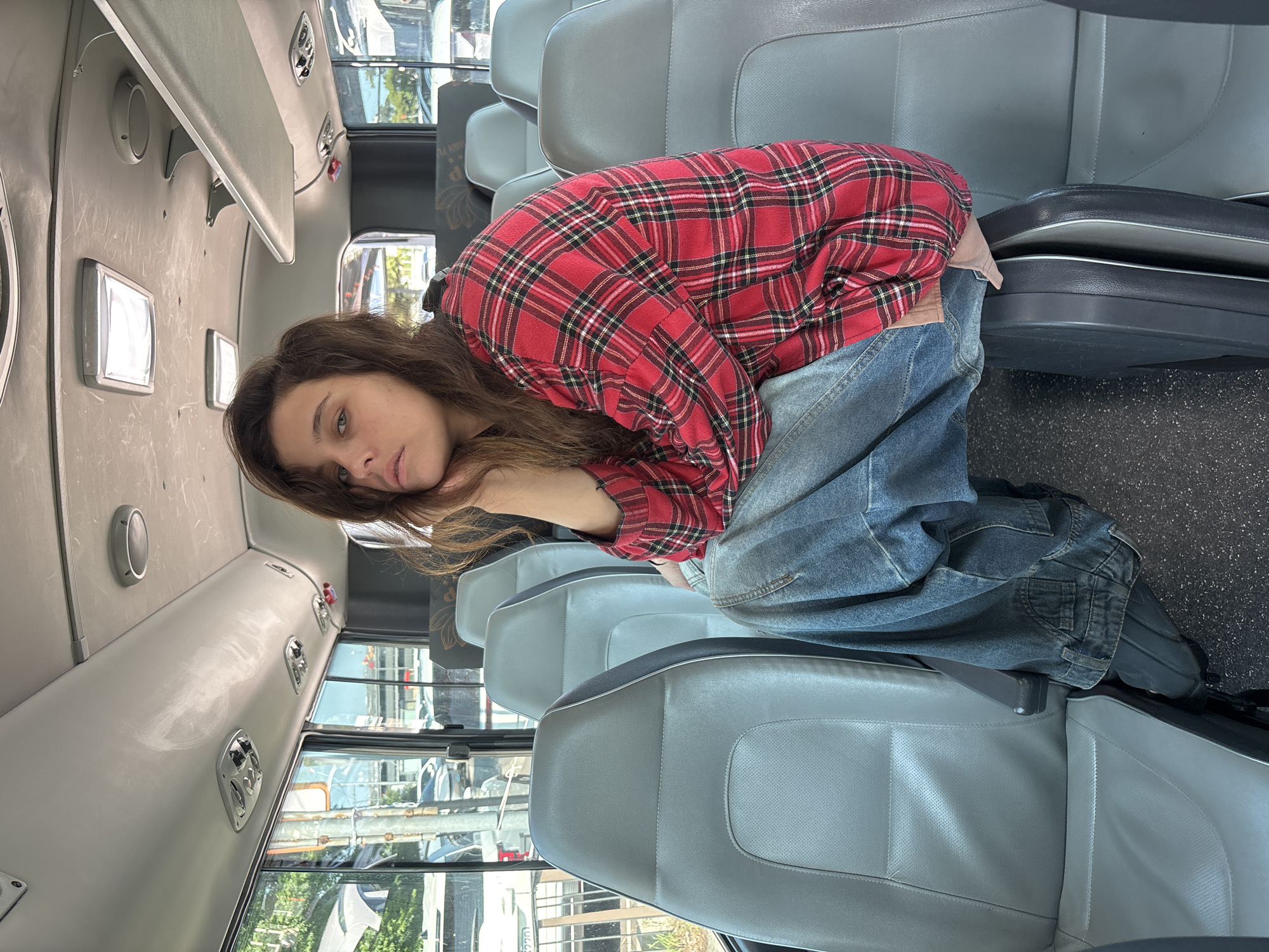 A young woman with long, brown hair is curled up on a bus seat, resting her head on her hands. She is wearing a red and black plaid shirt and baggy blue jeans. The bus has rows of gray seats with dark gray trim. The interior of the bus appears to be well-lit, with overhead lights visible. Through the windows, trees and other vehicles can be seen, suggesting it is daytime and possibly sunny. The overall mood is one of quiet introspection or fatigue. The location cues, such as the style of the bus and the surroundings visible through the windows, combined with the provided context of Jeju, South Korea, suggest the scene takes place on a local bus service. No visible text is present in the image.