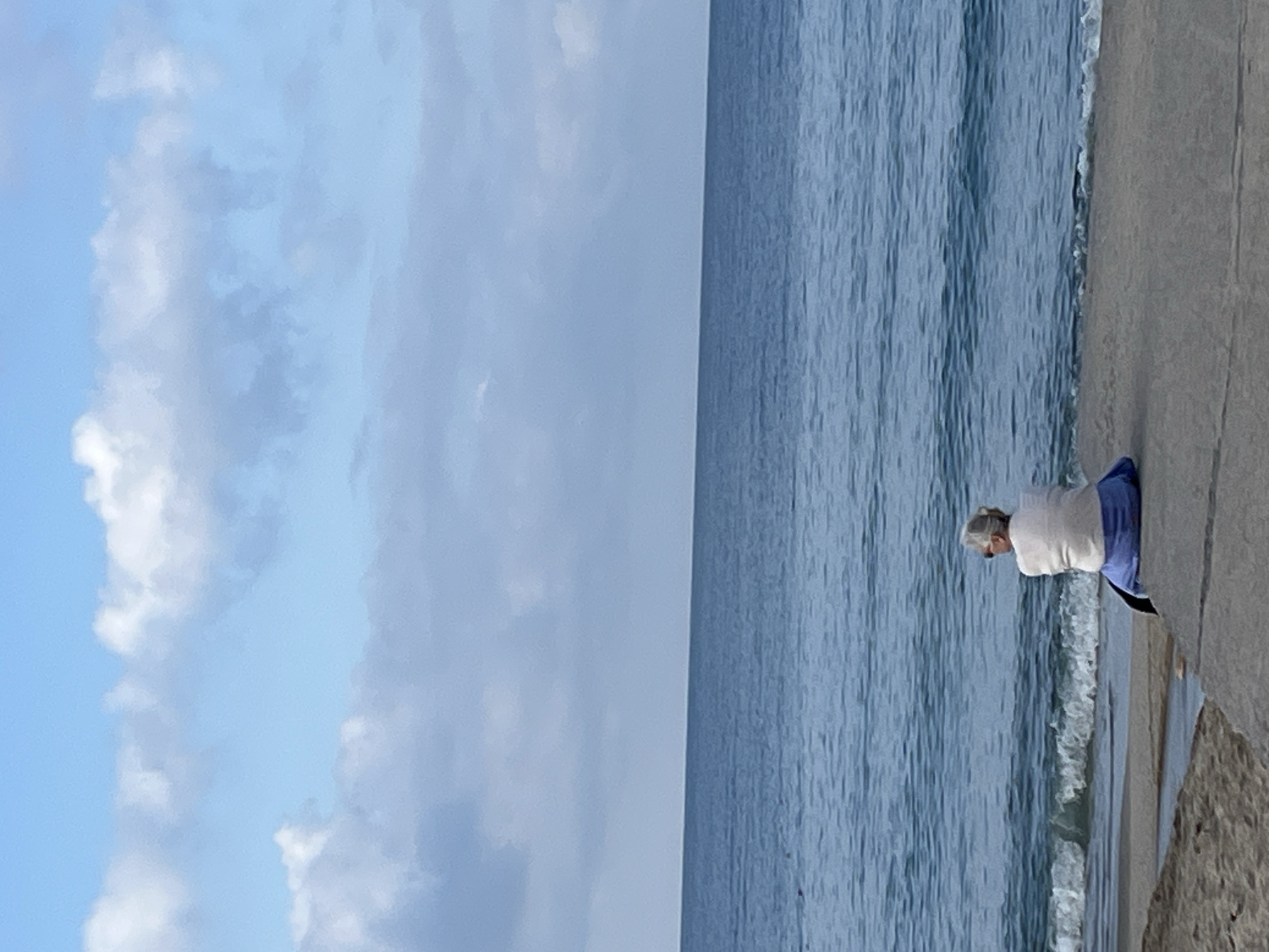 The image shows a view of the Mediterranean Sea in Alanya, Turkey. The sky is light blue with scattered white clouds. The sea is a darker blue with gentle waves approaching the shoreline. An elderly woman stands at the edge of a concrete seawall, looking out at the water. She is wearing a white top and blue bottoms. The photo appears to be taken during daylight hours.