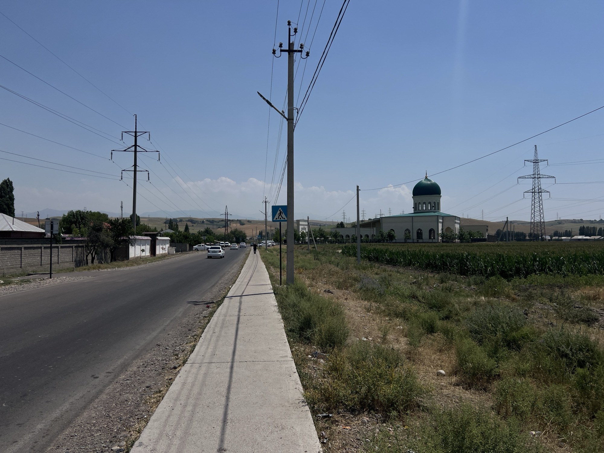 The image depicts a street scene in Karamanas, Uzbekistan. On the left side of the road, a gray wall and houses are visible, followed by a sidewalk. The road itself is asphalt, with several white cars driving on it. A lone pedestrian is walking on the sidewalk in the distance. To the right of the sidewalk, a field of green crops is present, with a large building featuring a green dome and arches in the background. Power lines and tall metal electricity towers are prominent features of the landscape throughout the image, with a clear blue sky overhead.