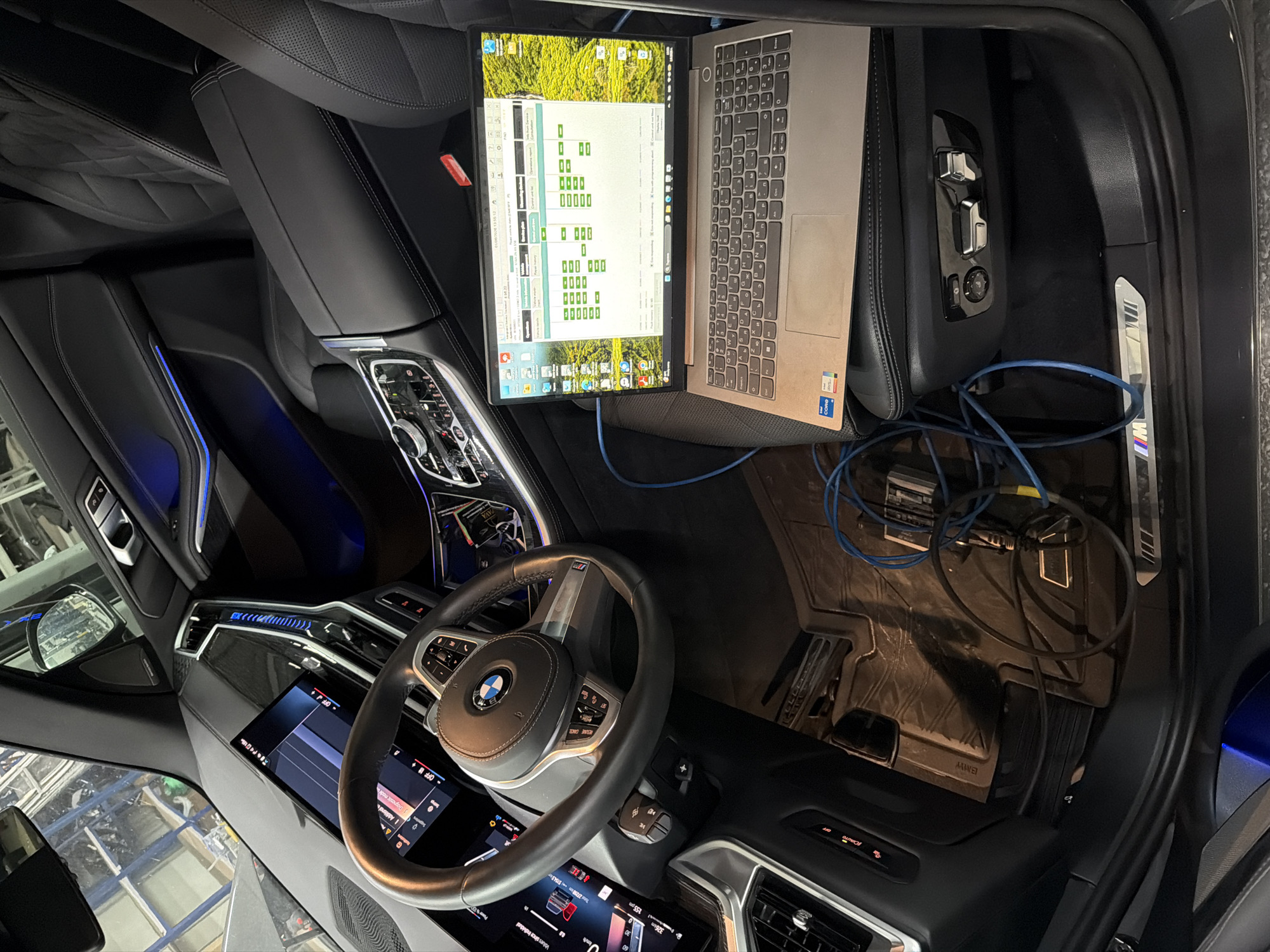 The image shows the interior of a modern BMW vehicle. A laptop is mounted on a stand near the passenger seat, displaying a diagnostic program. Cables connect the laptop to a device on the floor, suggesting a diagnostic or programming process is in progress. The interior features include leather seats, a digital dashboard, and modern styling elements such as ambient lighting. The image includes details like the BMW logo on the steering wheel, the "M" badge, and displays readouts like "326km" and "155 pm." The presence of diagnostic equipment suggests a repair or maintenance setting. The location, Burgas, Bulgaria, is not evident in the image.