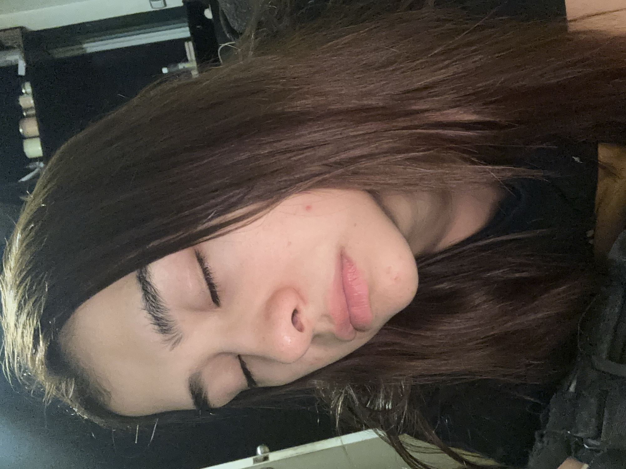 This media file is a close-up, slightly rotated portrait of a person sleeping or resting indoors. The image is rotated 90 degrees clockwise, depicting the person's head tilted to the right, implying they are lying on their left side.
The main subject is a person, likely female, with long, dark brown hair that partially covers their face and surrounds their head. Their eyes are closed, conveying a sense of peace and relaxation. Their facial features include natural, dark eyebrows and eyelashes, a discernible nose with visible pores, and slightly parted, full lips. A small, red blemish is visible on the person's forehead.
The individual appears to be wearing dark clothing, possibly a t-shirt or sweatshirt, visible around the neck and shoulder area. The background is dark and out of focus, suggesting an indoor setting like a bedroom or living space. Indistinct shapes in the upper left background, hinted at by the crops, suggest furniture or objects on a shelf.
The primary activity is sleeping or resting. No specific time of day is discernible from the lighting, but the posture and closed eyes strongly indicate a state of repose. There are no visible location cues within the image itself that relate to Puebla, Mexico, nor is there any visible text. The overall impression is one of calm and tranquility.