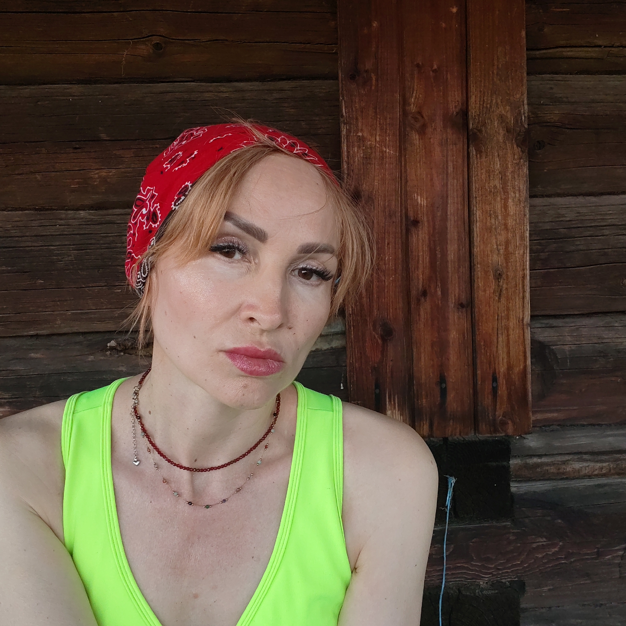 A fair-skinned woman with reddish-blonde hair is shown in a close-up shot, wearing a bright green tank top and a red bandana with a white paisley pattern on her head. She has two delicate necklaces around her neck. She has light eye makeup and her lips are lightly tinted. Her expression appears to be somewhat serious or contemplative.
The background consists of weathered, dark-brown wooden planks, indicating a rustic or outdoor setting, with two boards that appear to be newer. The location is in the city of Chaikovsky, Russia. A thin, blue cord is visible near the woman's shoulder in the background.