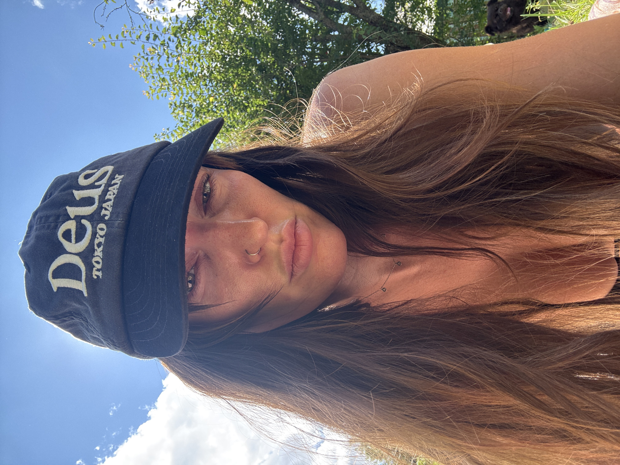 A woman with long brown hair and a black baseball cap that reads "Deus Tokyo Japan" is shown outdoors. She is wearing a necklace and has a nose ring. Sunlight illuminates her face and hair, and a tree is visible in the background, along with a black dog. The image appears to be a selfie taken on a sunny day, possibly in a park or backyard setting. The location is identified as Krasnaya Presnya SNT, Russia.