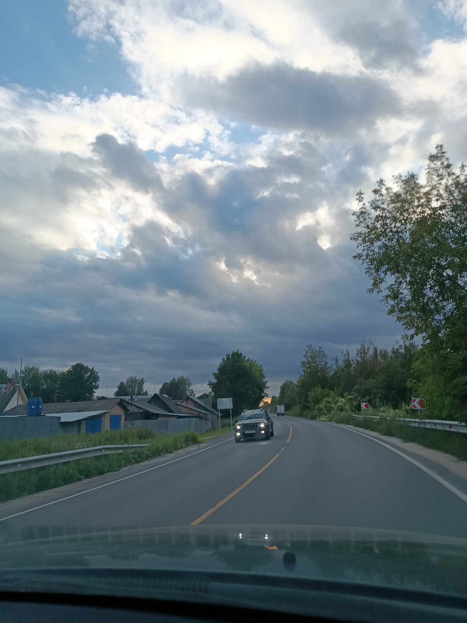 The image depicts a view from the interior of a vehicle traveling on a road, likely in or near the village of Snopok Staryy, Russia. The road curves to the right and is marked with a yellow line. Ahead, a dark-colored SUV with its headlights on is visible in the left lane, and further down the road, a white truck is also traveling in the same direction. The right side of the road is bordered by a metal guardrail and dense green trees. On the left side of the road, there are several low-rise buildings with gray and tiled roofs, some appearing to be houses or sheds, situated behind a fence. The sky above is filled with dramatic, dark gray clouds with patches of bright sky visible through them, suggesting overcast or potentially stormy weather. The overall lighting suggests it is late afternoon or early evening.