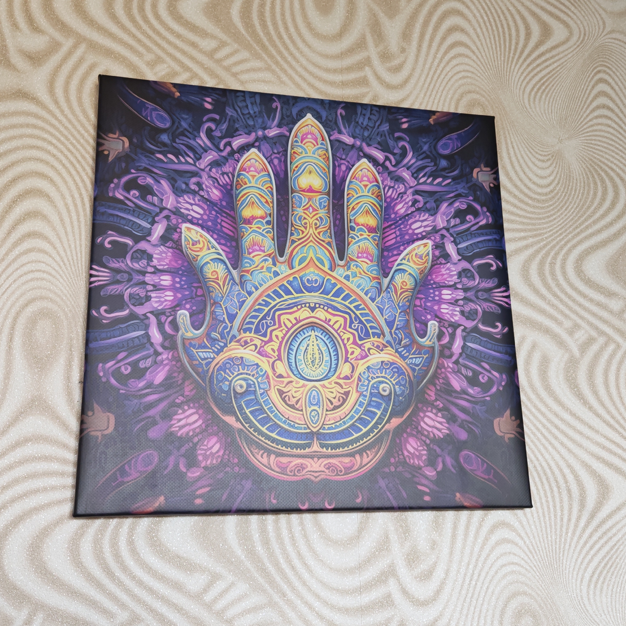 A square canvas print featuring a stylized Hamsa hand with an eye in the center is displayed against a background with swirling, wavy lines. The Hamsa hand is intricately designed with various colorful patterns and shapes in shades of yellow, blue, purple, and pink. The background of the print is dark with purple floral patterns. The print appears to be resting or mounted against a wall. The wavy lines behind the canvas are shades of beige. There are no people visible in the image. The location is not specified as Rostov, Russia and there is no clear information on location from this image.