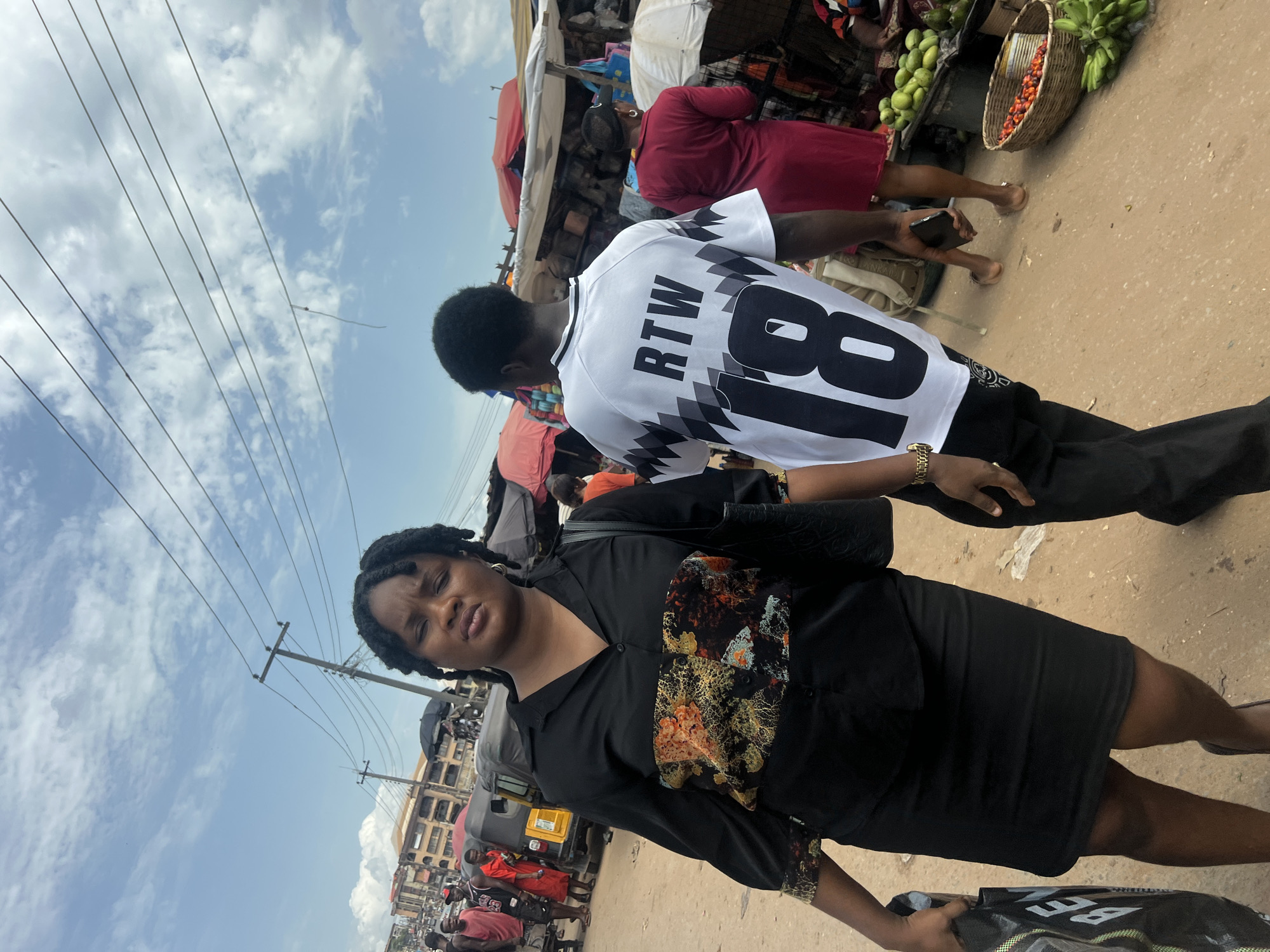 The image depicts two individuals in a bustling city market environment in Unknown, Unknown. In the foreground, a woman is visible, wearing a black dress with a patterned floral design around the midriff. She appears to be holding a bag in her right hand. Behind her and slightly to the right, a man is wearing a white jersey with the letters "RTW" and the number "18" printed on the back. The market is crowded with people and stalls. Visible items include fresh produce in baskets and beneath umbrellas. Overhead, power lines are visible against a sky with scattered clouds.