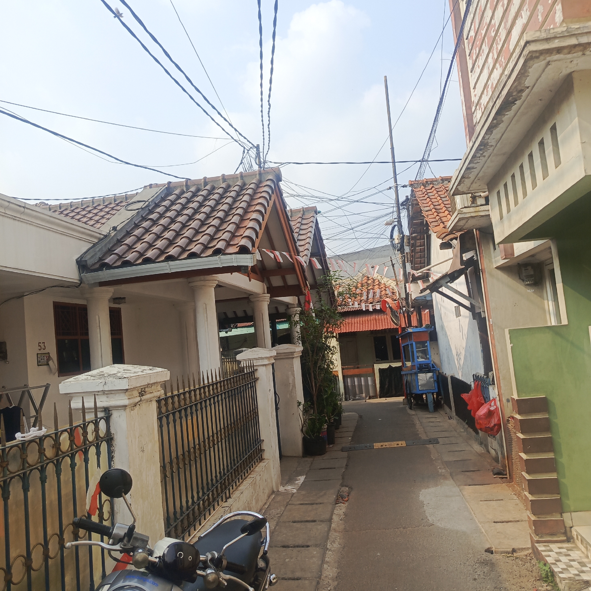 The image depicts a narrow street in a residential area, possibly in South Jakarta, Indonesia, characterized by a mix of houses and overhead electrical wires. The sky is visible and appears to be daytime. On the left side of the street, a white house with a tiled roof is partially visible, featuring a black wrought-iron fence and a white pillar. A motorcycle is parked in the foreground, facing towards the viewer, with its handlebars and front portion clearly visible.
The street itself is paved with asphalt and has a concrete drainage channel running alongside it. Several potted plants are placed along the sidewalk. On the right side of the street, another building with a green facade and a beige upper section is visible, with a set of outdoor stairs leading up to it. Further down the street, a small blue and orange cart, possibly a food stall, is parked. Red plastic bags are hanging on a wall near the cart.
The overall impression is of a densely populated urban environment with visible signs of everyday life, including residential structures, infrastructure, and street vendors. The presence of numerous electrical wires crisscrossing the sky is a notable feature. The image seems to capture a moment of ordinary street life in a South Jakarta neighborhood.